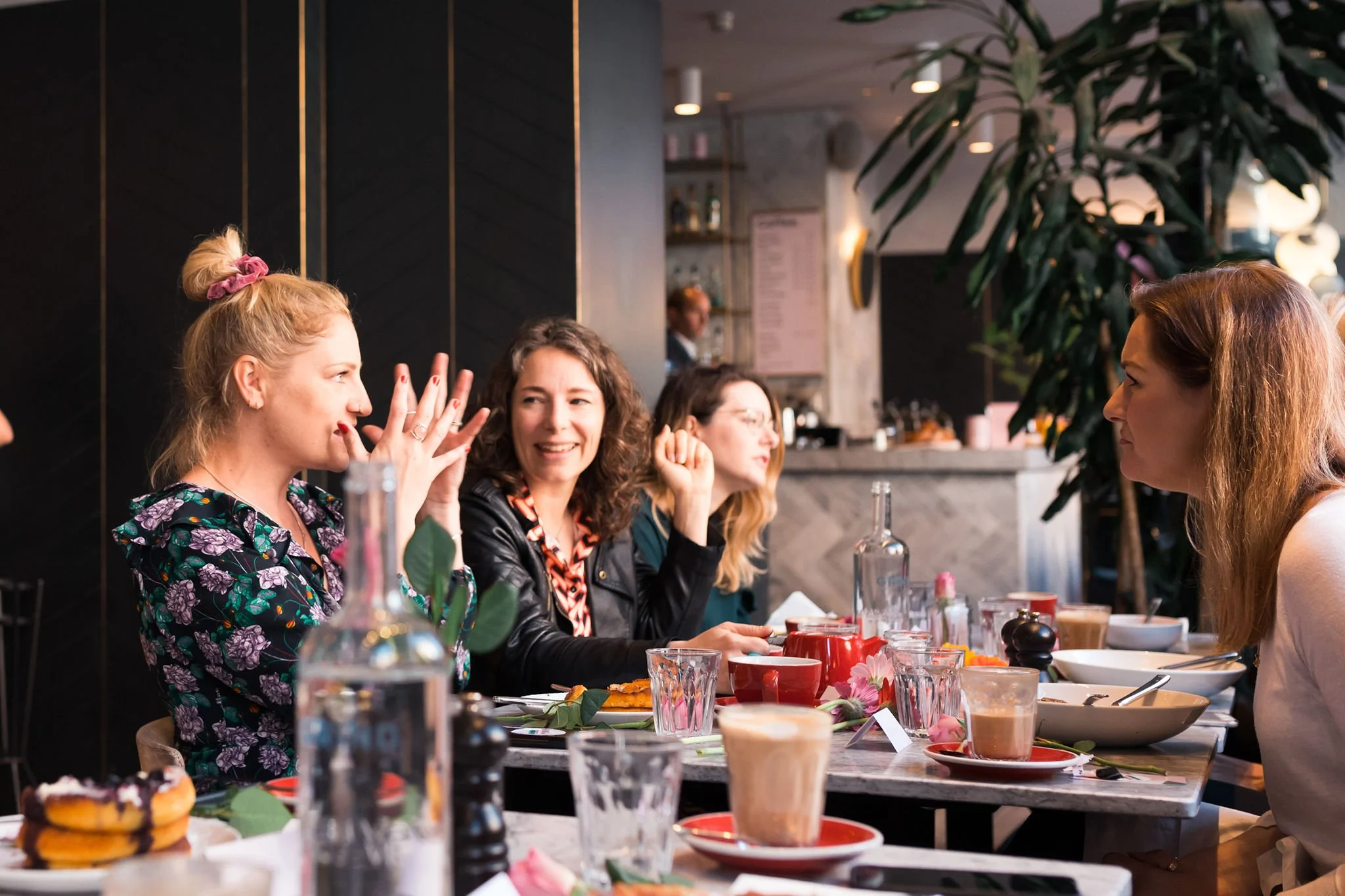 A group of four women sitting at a restaurant table, engaging in conversation with food and drinks on the table.