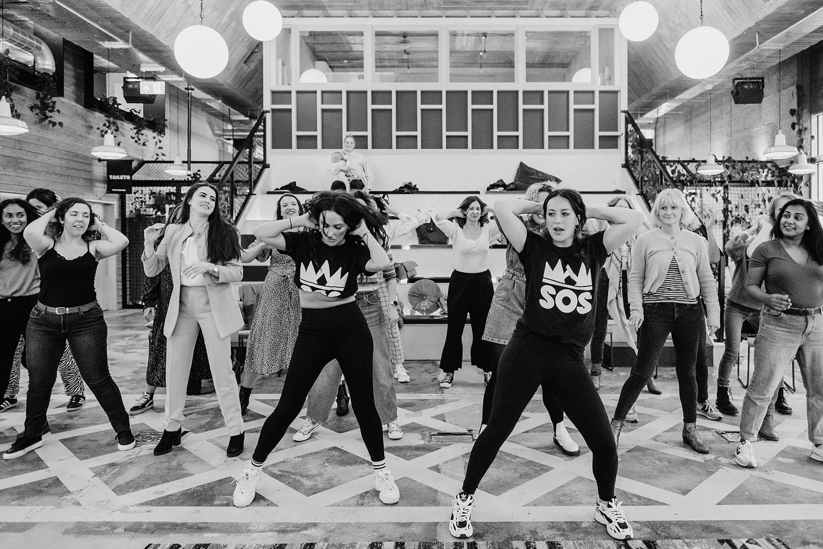 A group of women dancing and practicing choreography inside a modern, well-lit venue with wooden accents and hanging spherical lights.