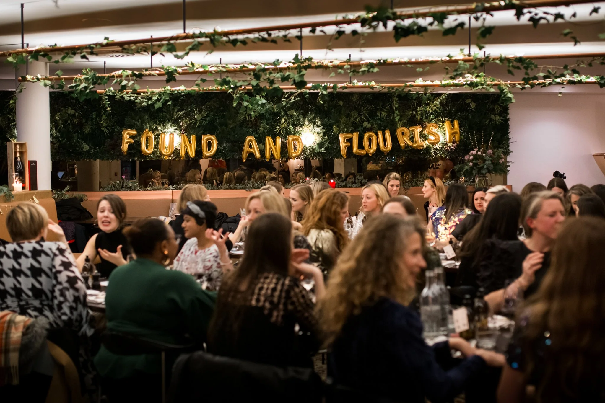 A crowded indoor event with many women seated at tables, decorated with a greenery backdrop and gold balloon letters spelling 'FOUND AND FLOURISH'