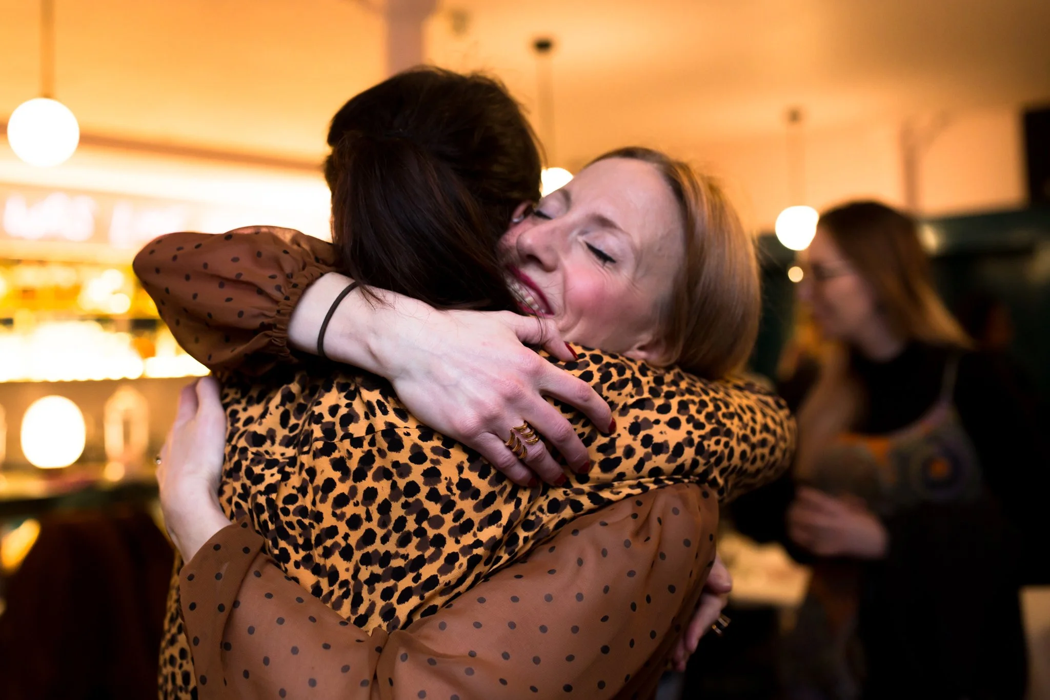 Two women hugging warmly, smiling with closed eyes, in a cozy indoor setting with warm lighting.
