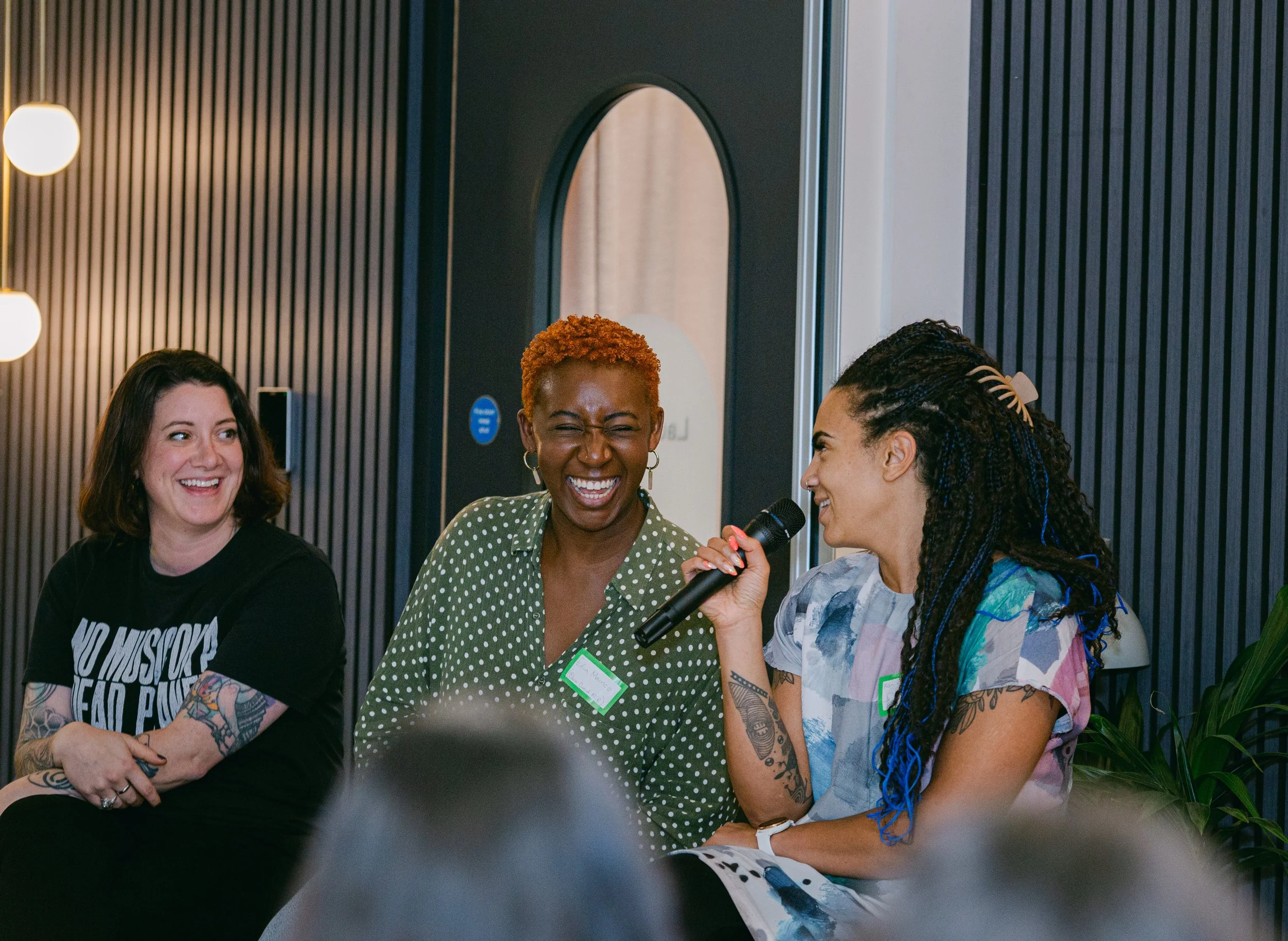 Three women sitting on a panel, with one woman holding a microphone and speaking, all smiling and laughing during a discussion.