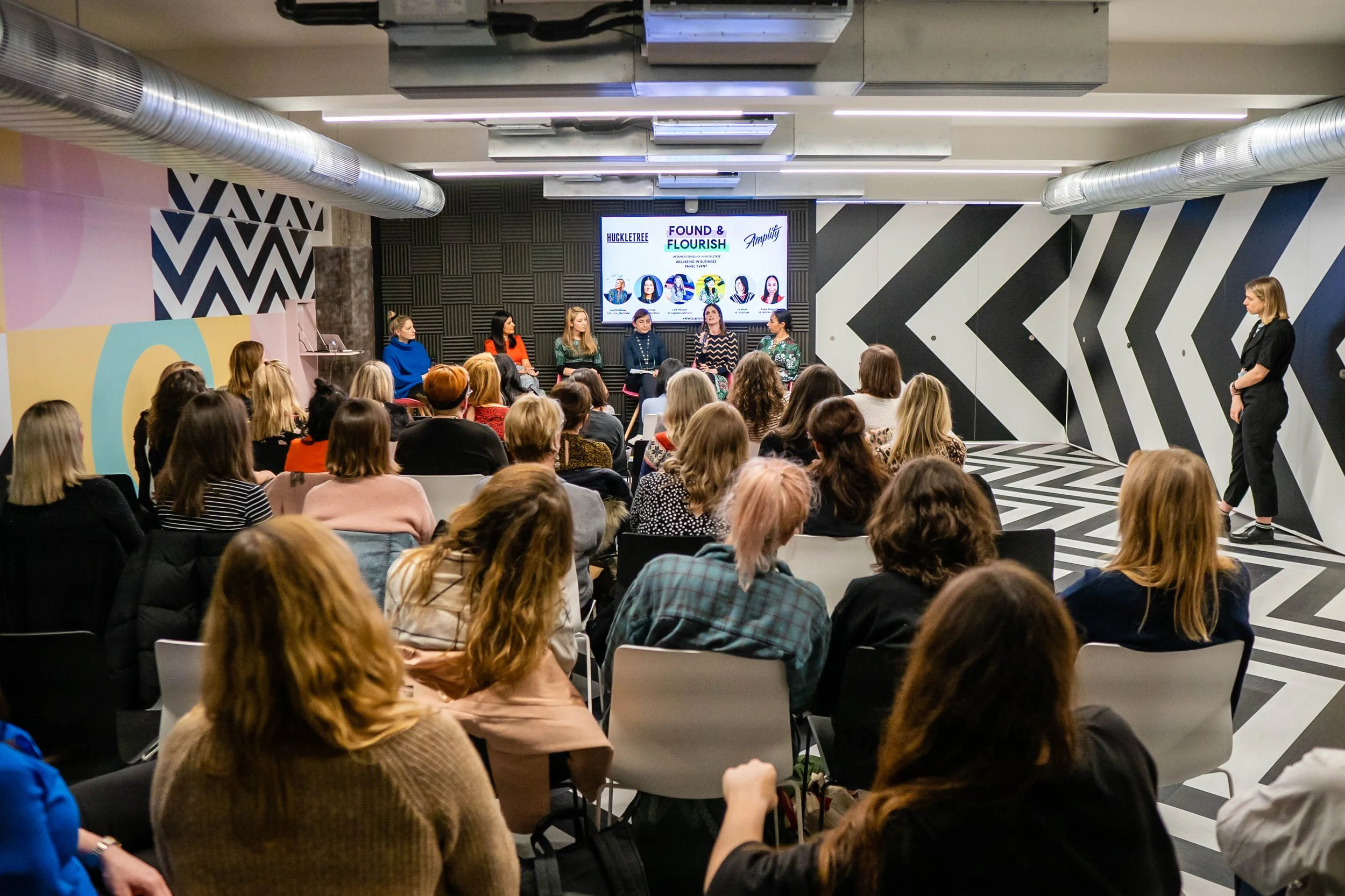 A conference or panel discussion with multiple speakers seated at the front of a room filled with seated audience members, with a large digital screen behind the speakers displaying event details.