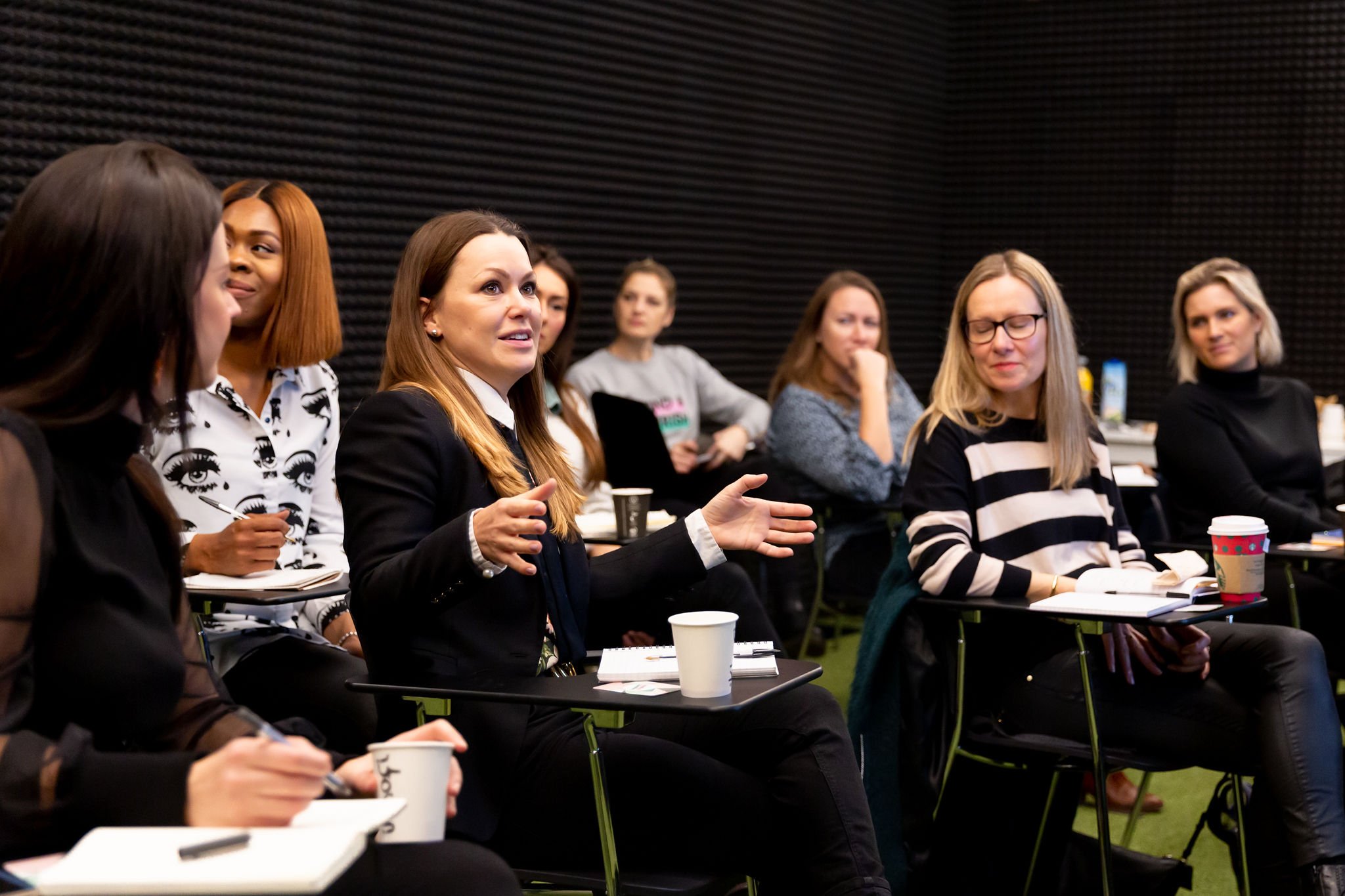 Group of women attending a discussion or workshop in a room with black acoustic panels, some taking notes, and a woman in a blazer speaking gesturing with her hands.