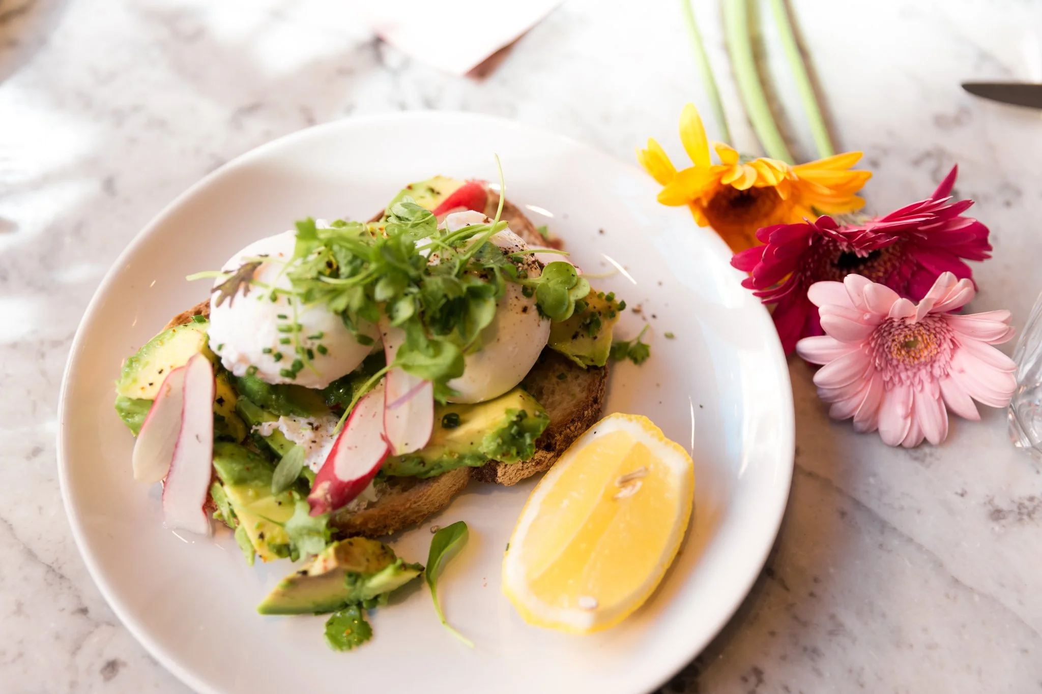 Poached eggs on toast with avocado, radish slices, microgreens, and a lemon wedge, served on a white plate with colorful flowers nearby.