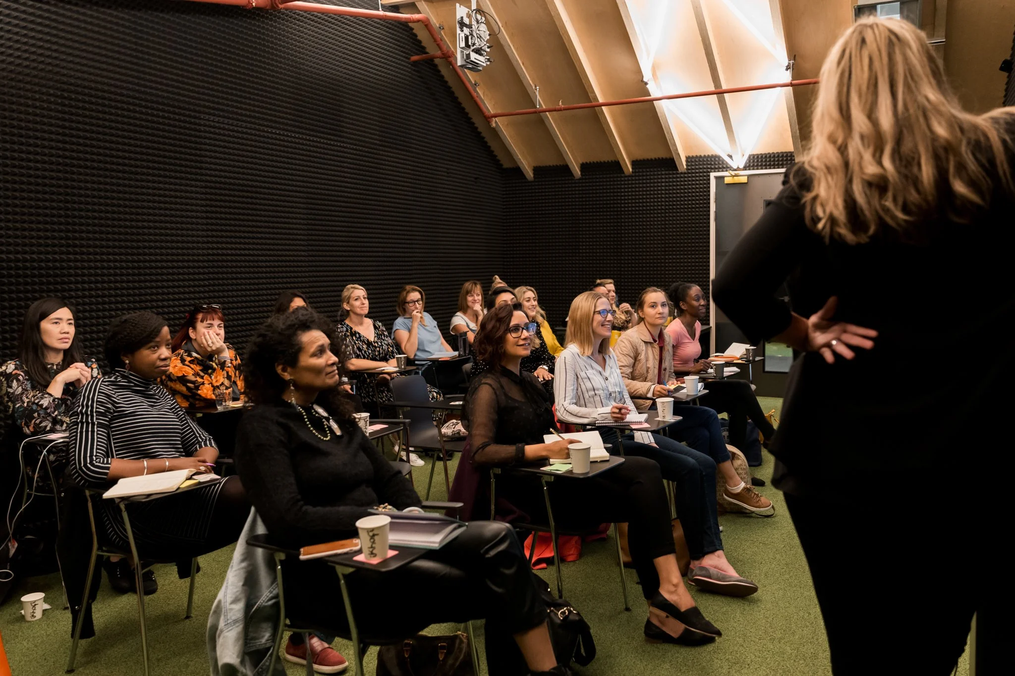 A group of women attending a seminar or workshop, seated in rows with notebooks and cups, listening to a speaker standing at the front of the room.