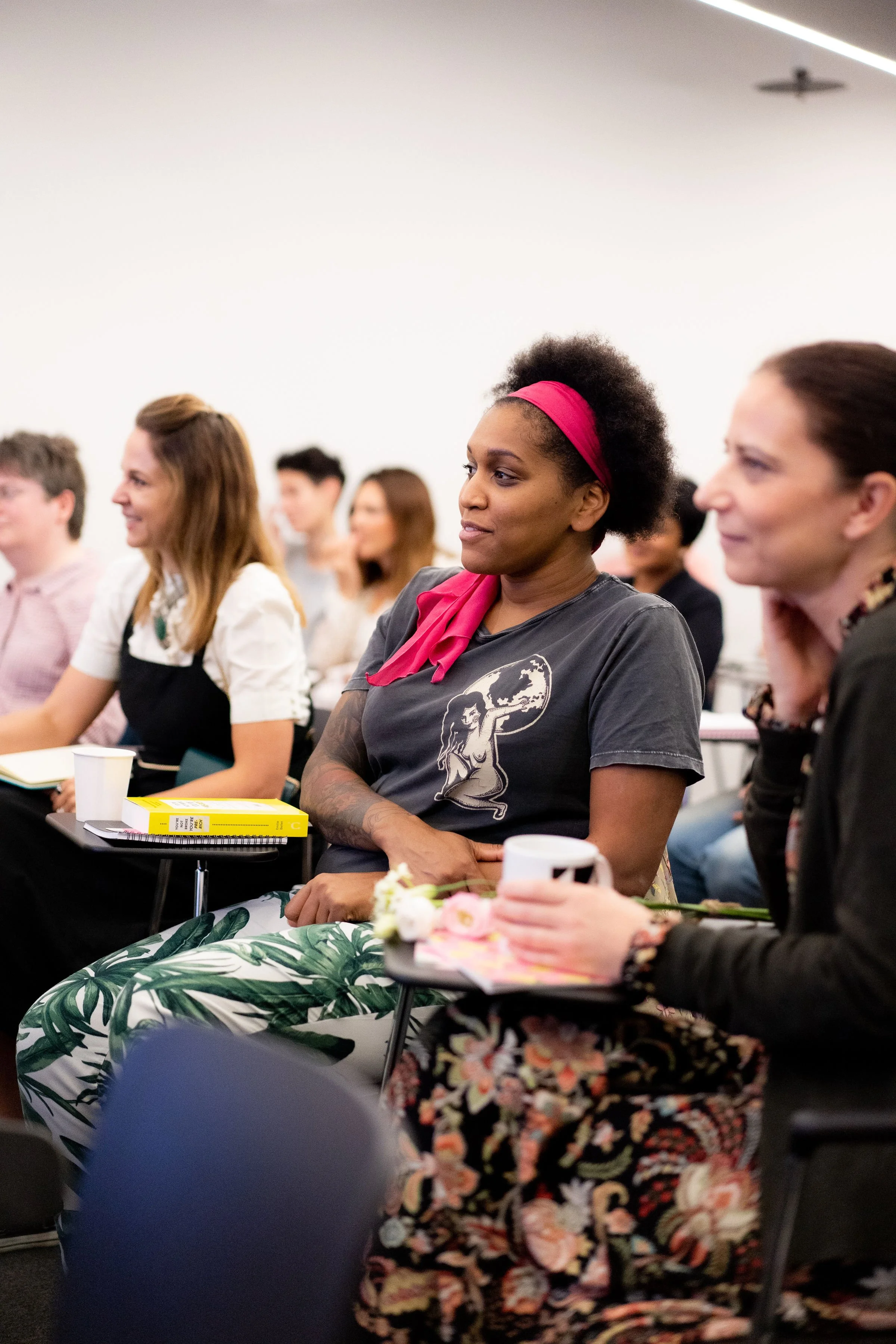A diverse group of people, mostly women, sitting and attentively listening at a seminar or conference in a well-lit room.