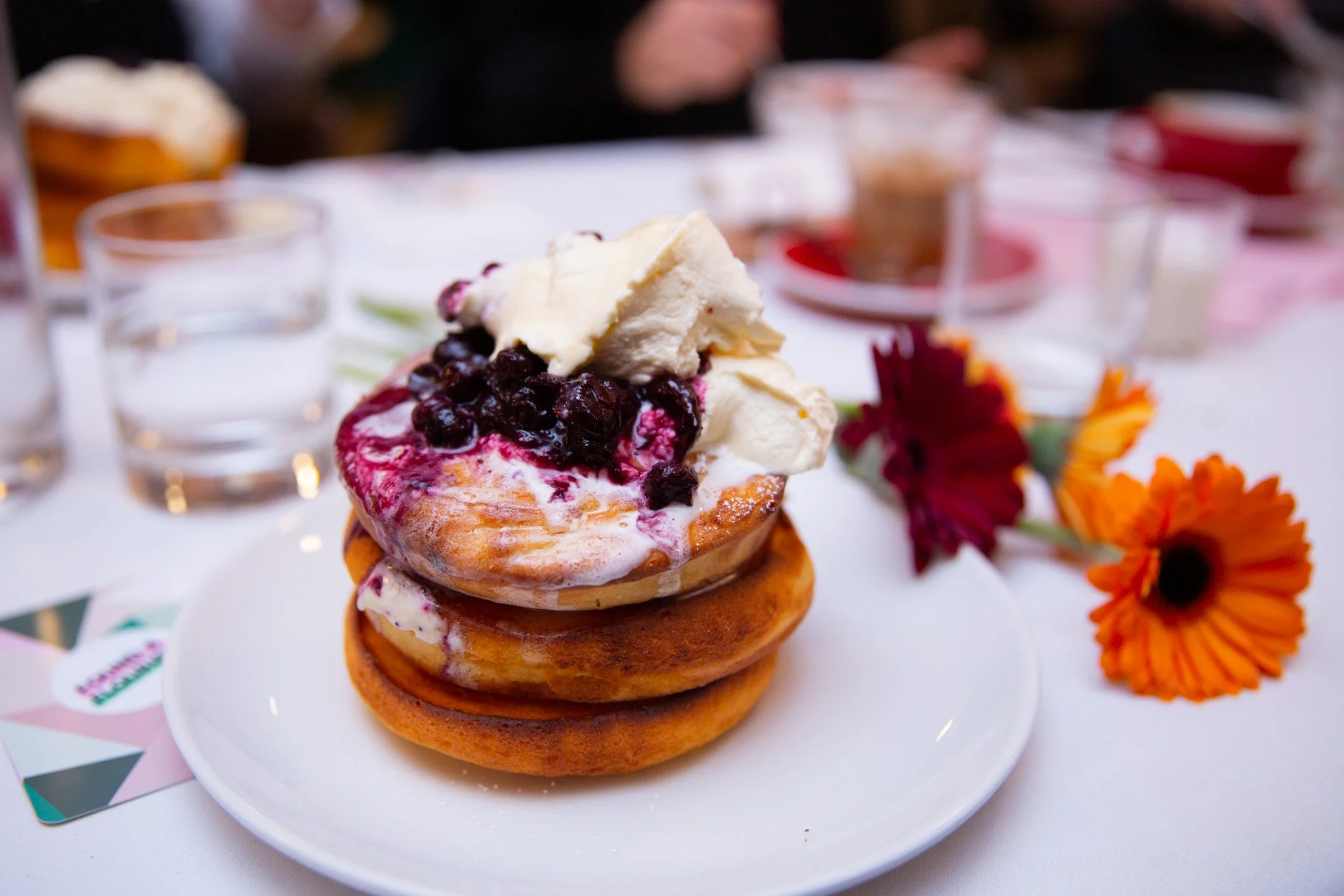 Stack of two golden-brown waffles topped with vanilla ice cream and blueberry compote on a white plate, with orange and red flowers in the background.