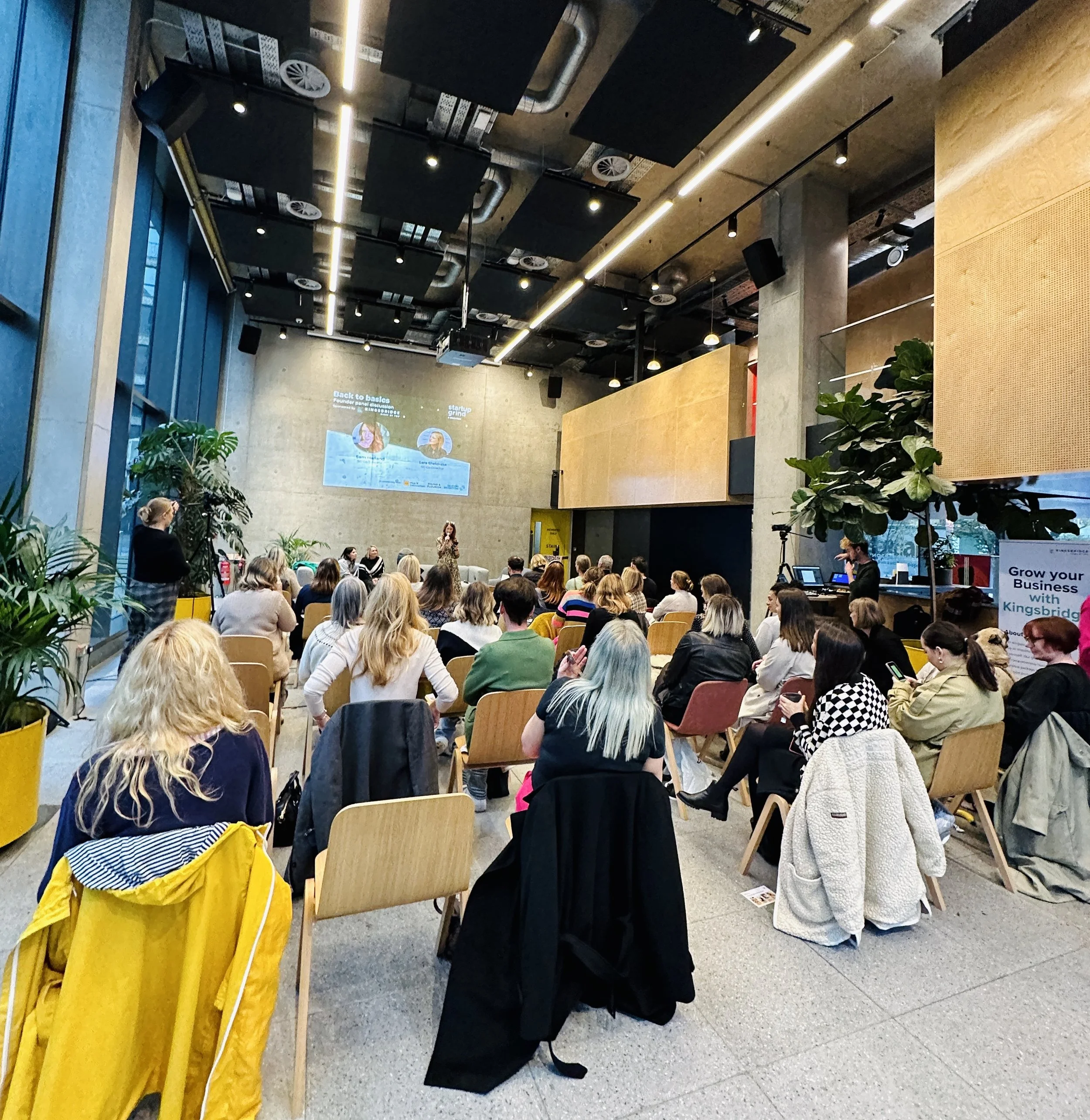 A conference or seminar room filled with people seated in chairs, facing a speaker at the front. There is a large screen displaying a presentation, potted plants on the sides, and a photographer in the corner.