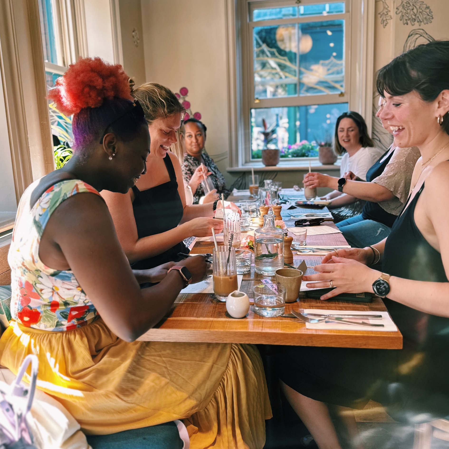 A group of women sitting at a restaurant table, enjoying drinks and conversation, with windows and decorative plants in the background.