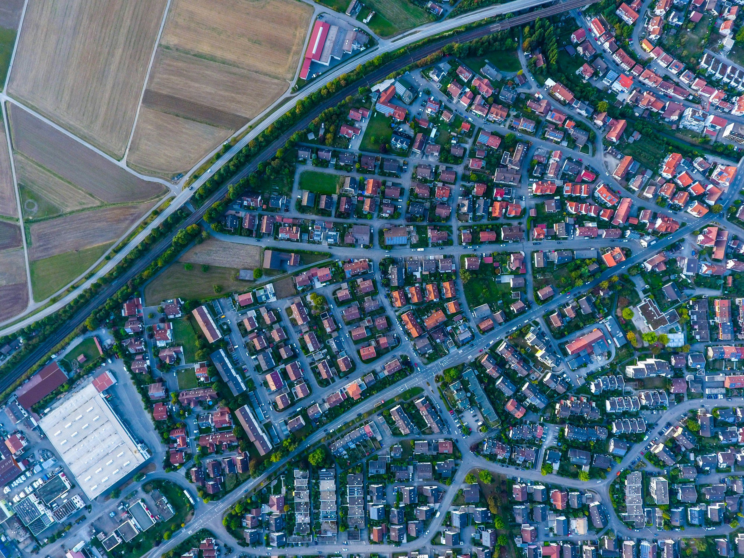 An aerial view of a suburban neighborhood with houses, streets, and greenery, adjacent to open farmland and fields.