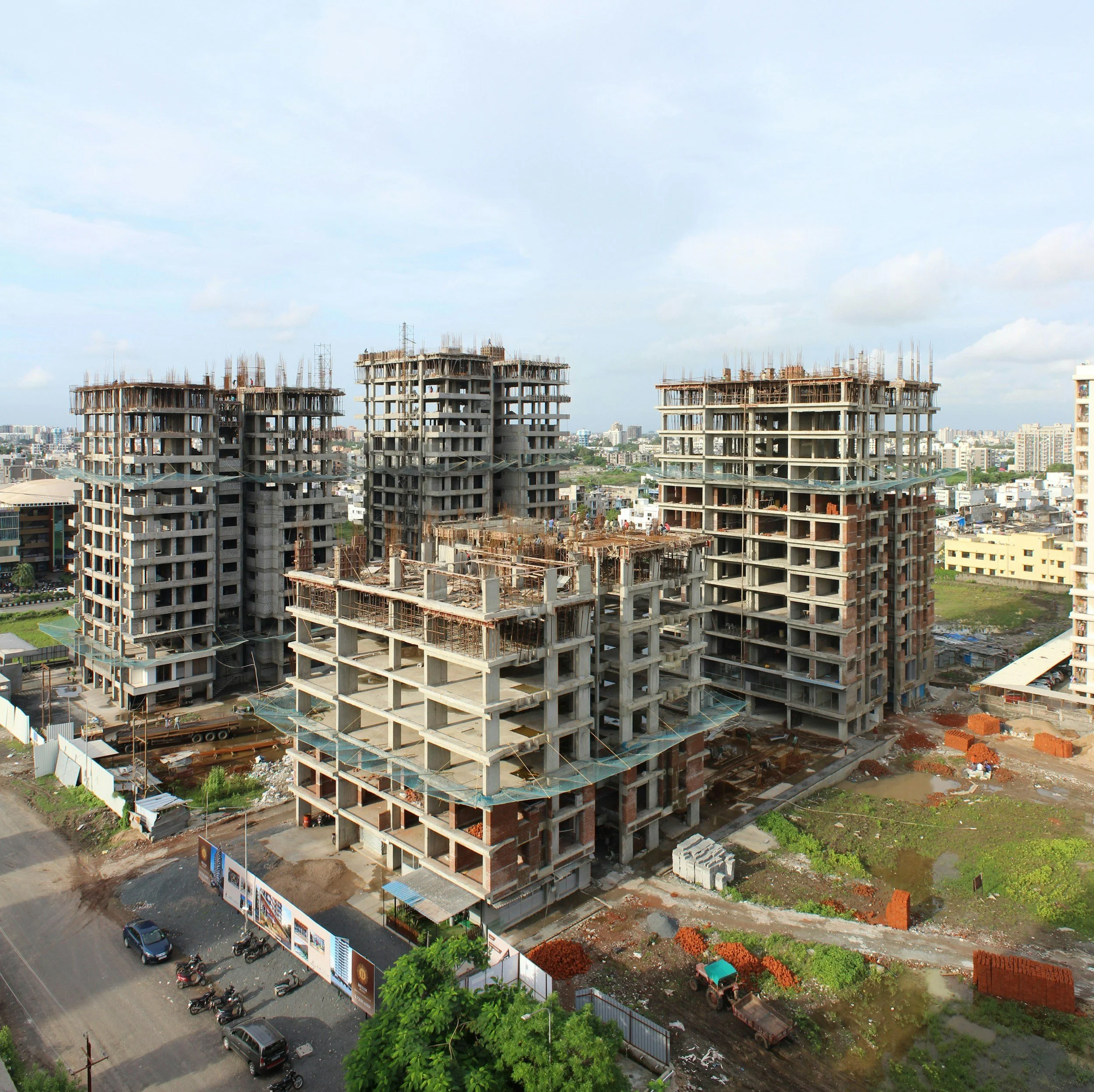 Construction site with multiple unfinished multi-story buildings in an urban area, surrounded by cityscape, roads with parked bikes and cars, and construction materials on the ground.