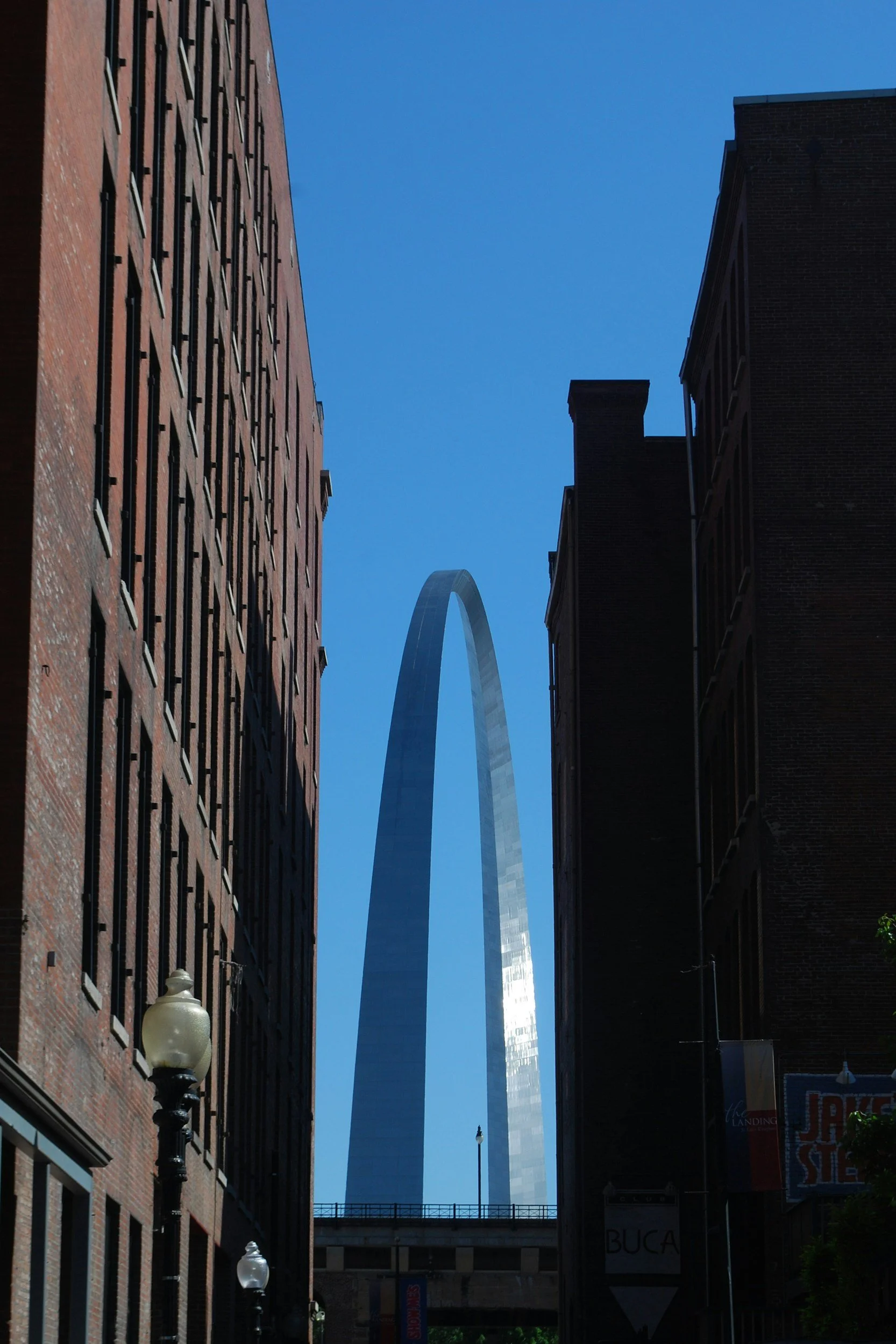 Photo taken from a street level, looking upward between two brick buildings with a bright blue sky in the background. The Gateway Arch in St. Louis, Missouri, is visible in the distance.