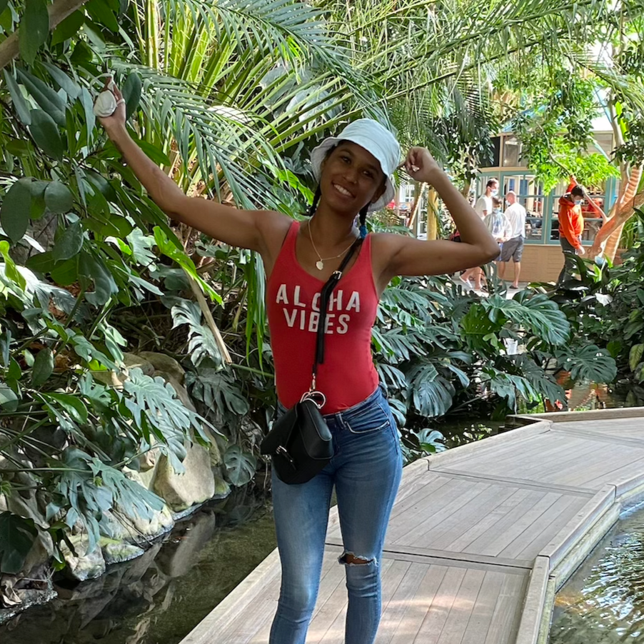 Femme souriante portant un t-shirt rouge avec l'inscription "Aloha Vibes", jeans, un chapeau blanc et une sacoche noire, debout sur une passerelle en bois dans un environnement tropical luxuriant.