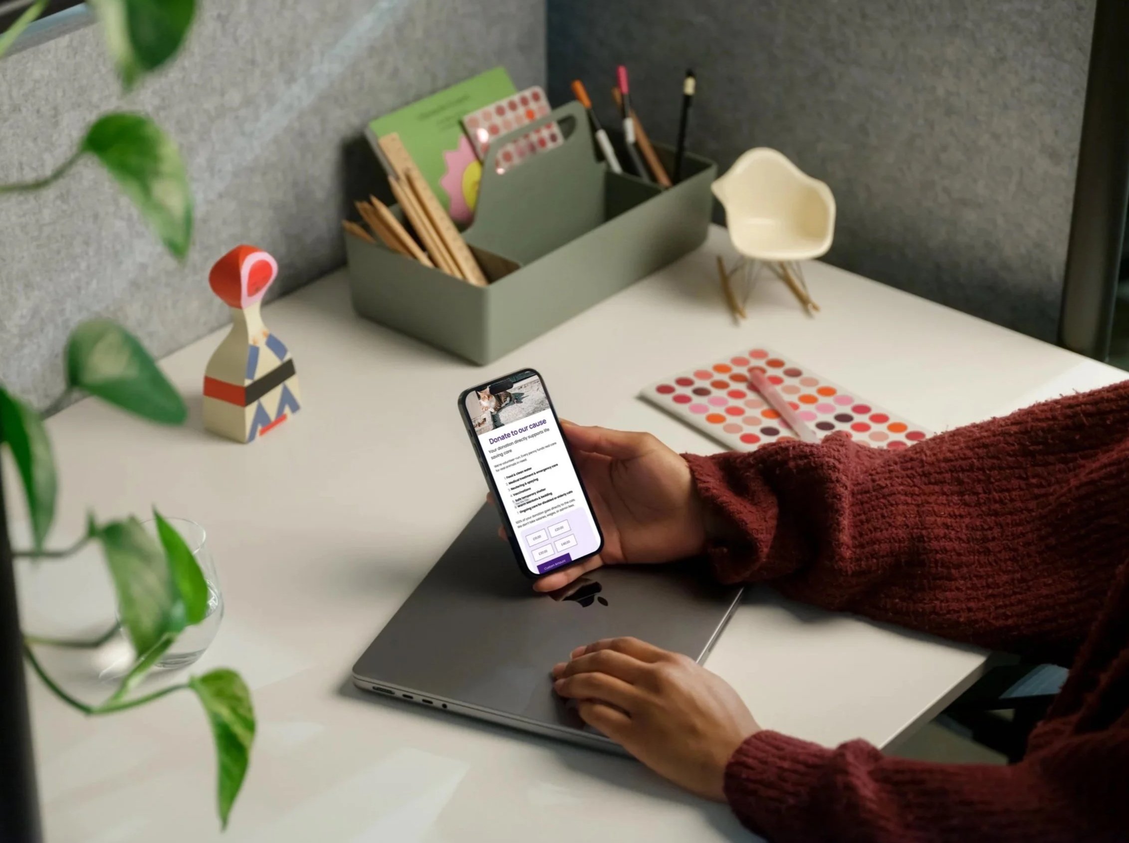 Person in a maroon sweater using a smartphone to make a donation at a desk with office supplies, a laptop, and a colorful palette.