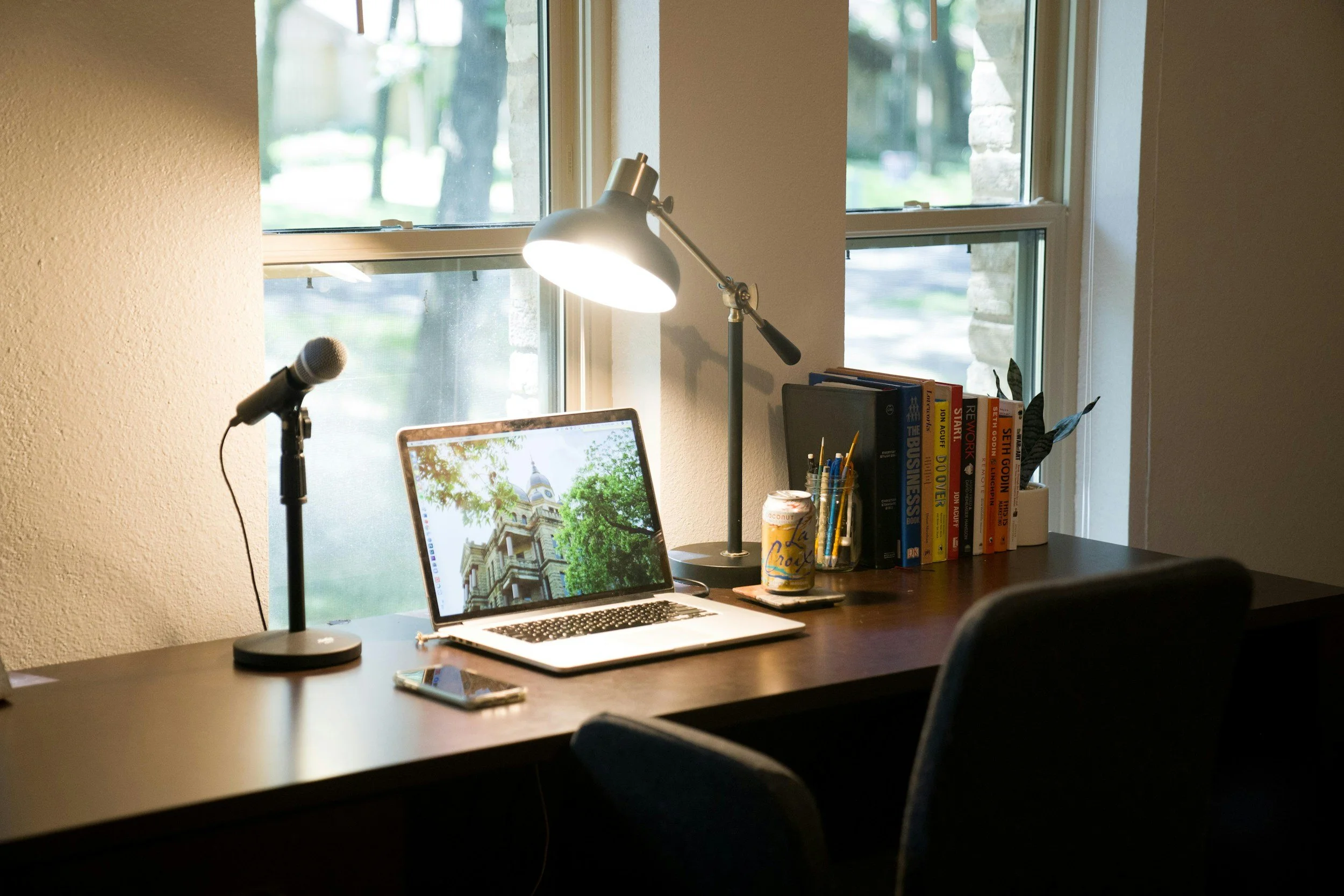 A home office desk with a laptop displaying a cathedral, a microphone on a stand, a desk lamp, a can of LaCroix soda, a smartphone, and a row of books, positioned in front of a window with a view of trees.