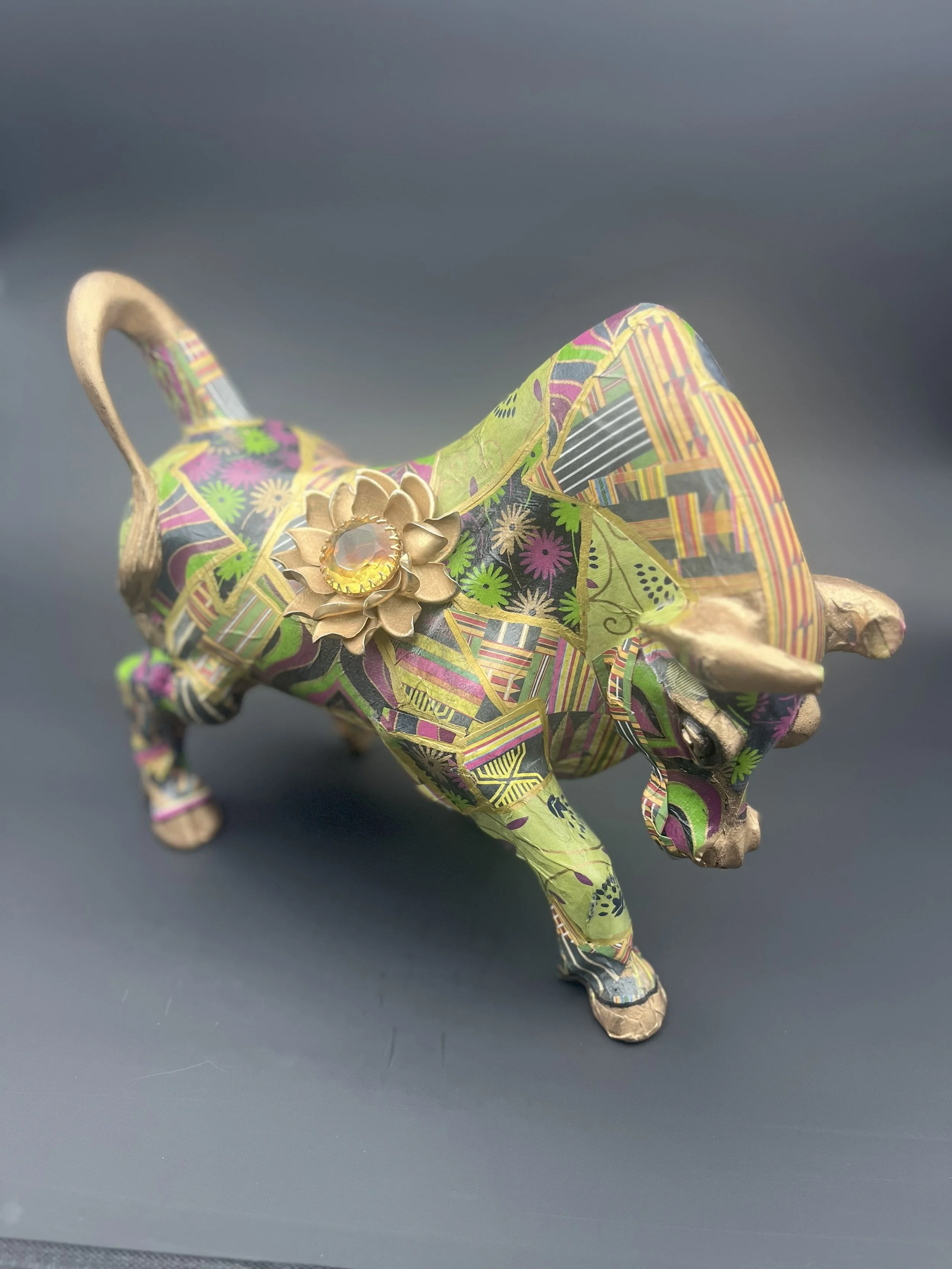 Bull in a Jewelry Shop
Artist: Vincent Dion Stringer
Year: 2024
Medium: Mixed Media Sculpture
Materials: Decoupage with Kente Cloth Paper, Gold Leaf, Vintage Brooch
Dimensions: [Insert Dimensions]

Description:A stylized charging bull, captured mid-s