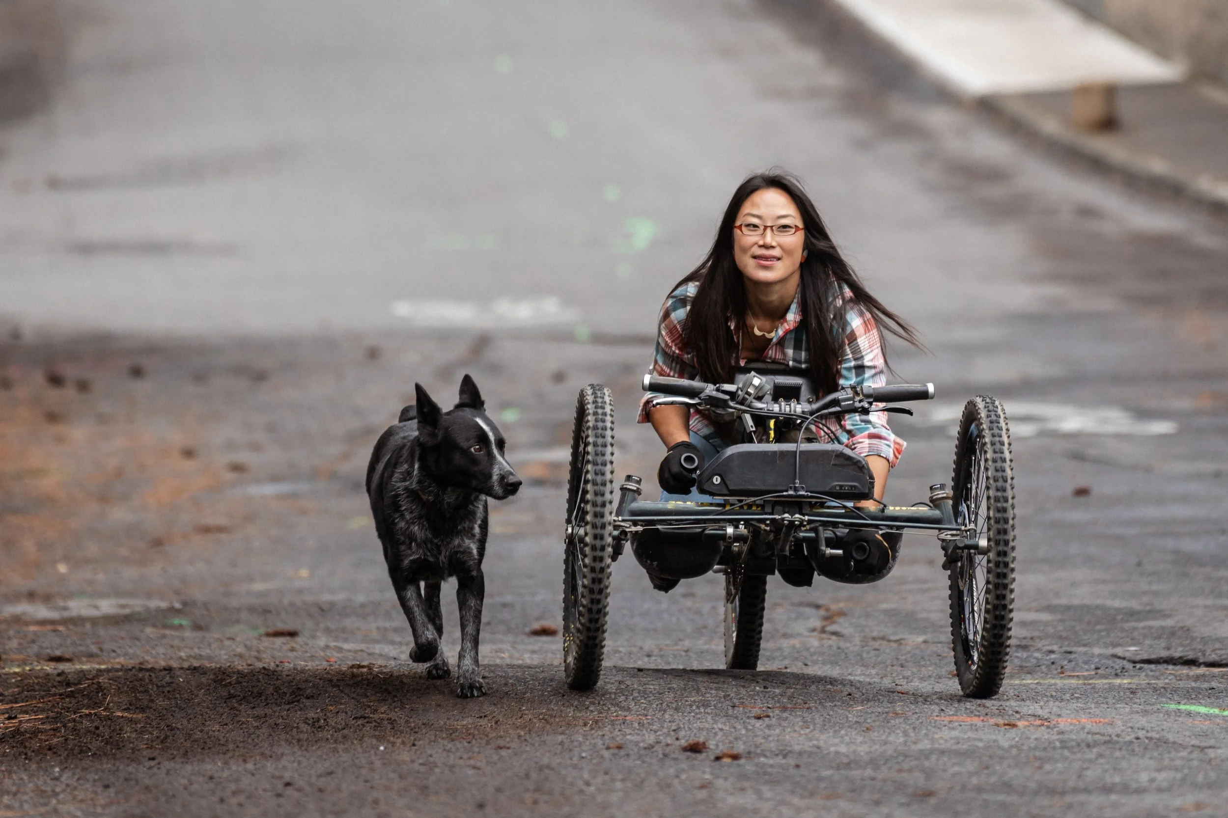 A woman with long dark hair and glasses sitting on a wheelchair with large tires, on a wet street, with a black and white dog walking beside her.