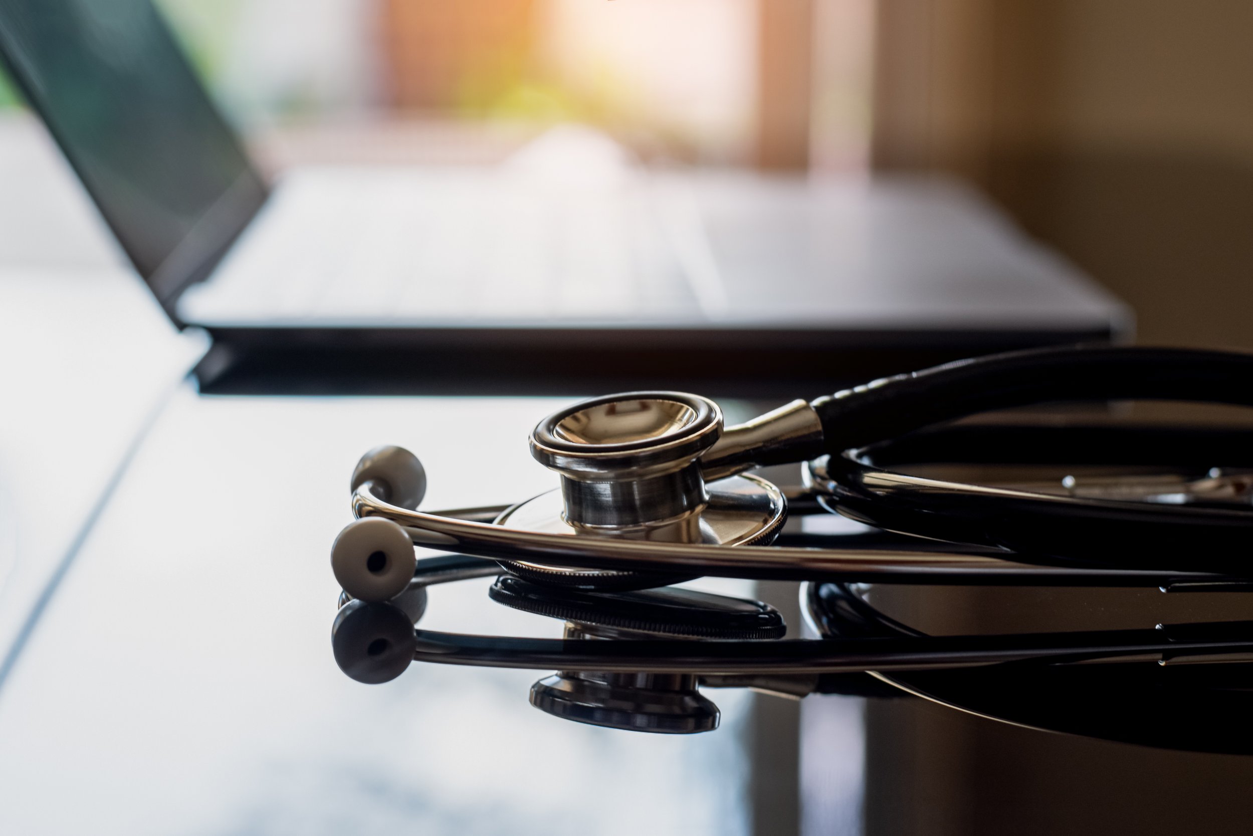 A stethoscope with black tubing and silver chest piece resting on a reflective black surface, with a laptop in the background.
