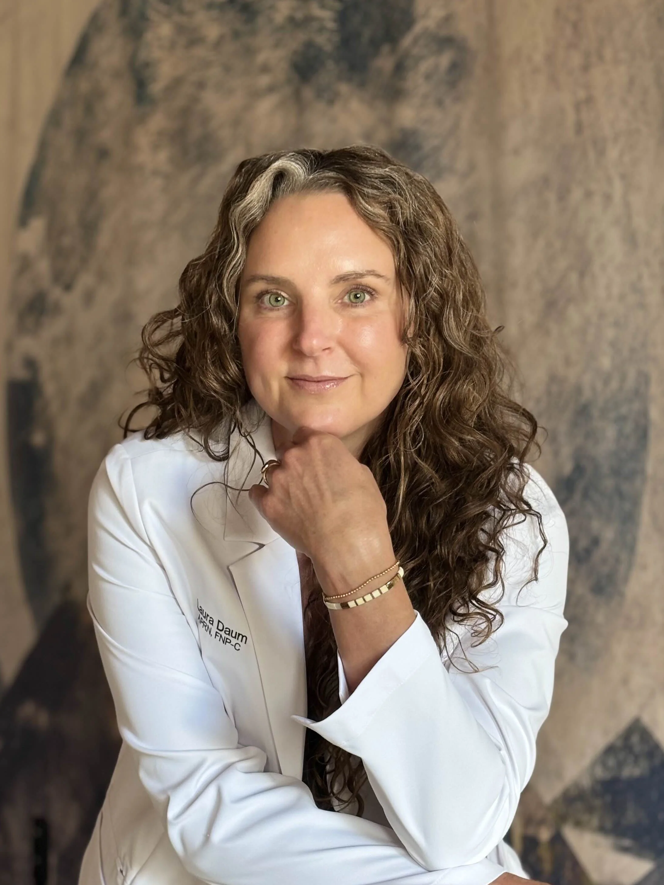 A woman with curly brown hair and green eyes, wearing a white medical coat with name tag, sitting with her hand resting under her chin, looking at the camera, against a blurred brown and gray background.