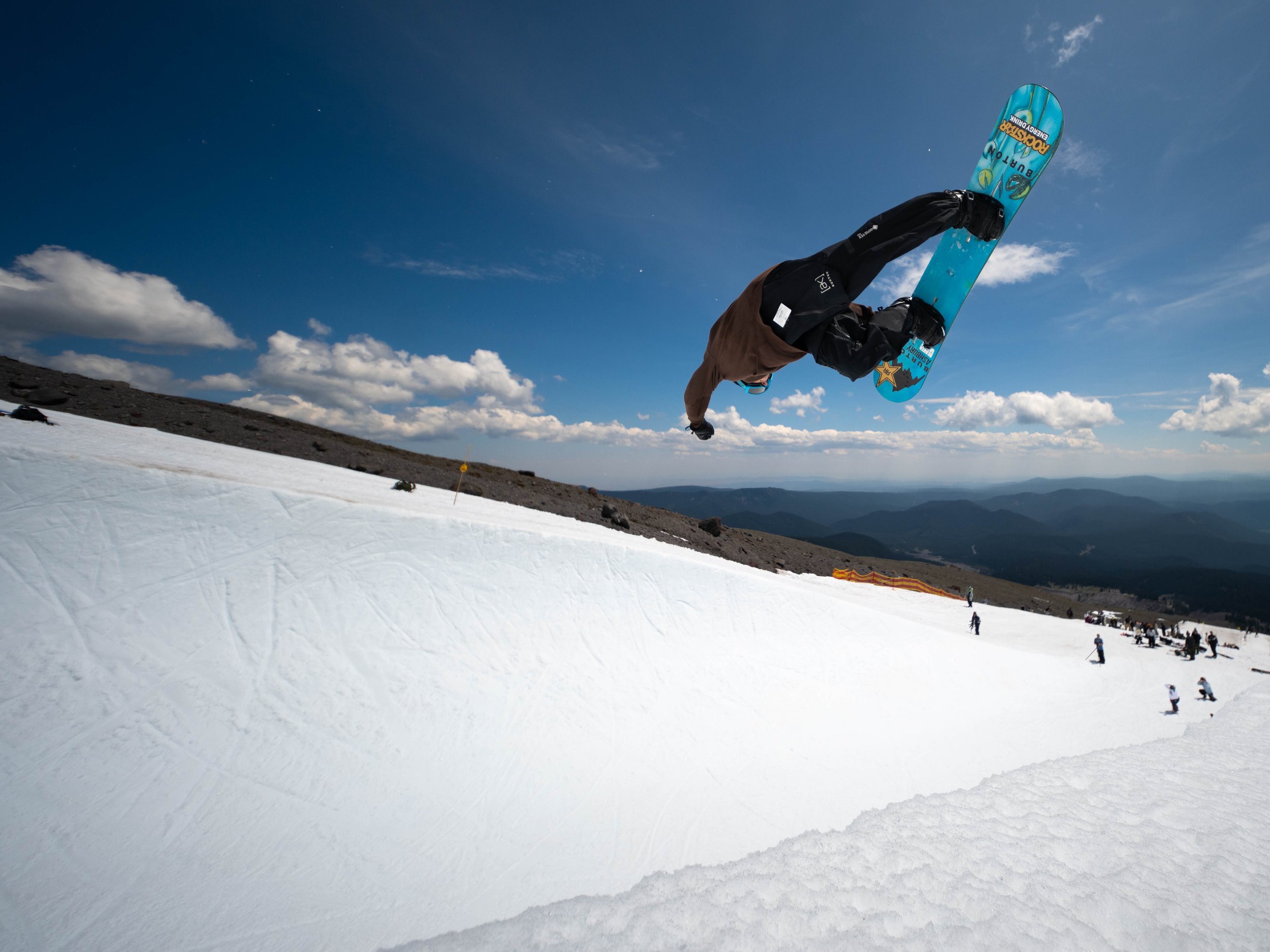 Snowboarder performing a grab in mid-air on a snowy slope with a mountain landscape and blue sky in the background.