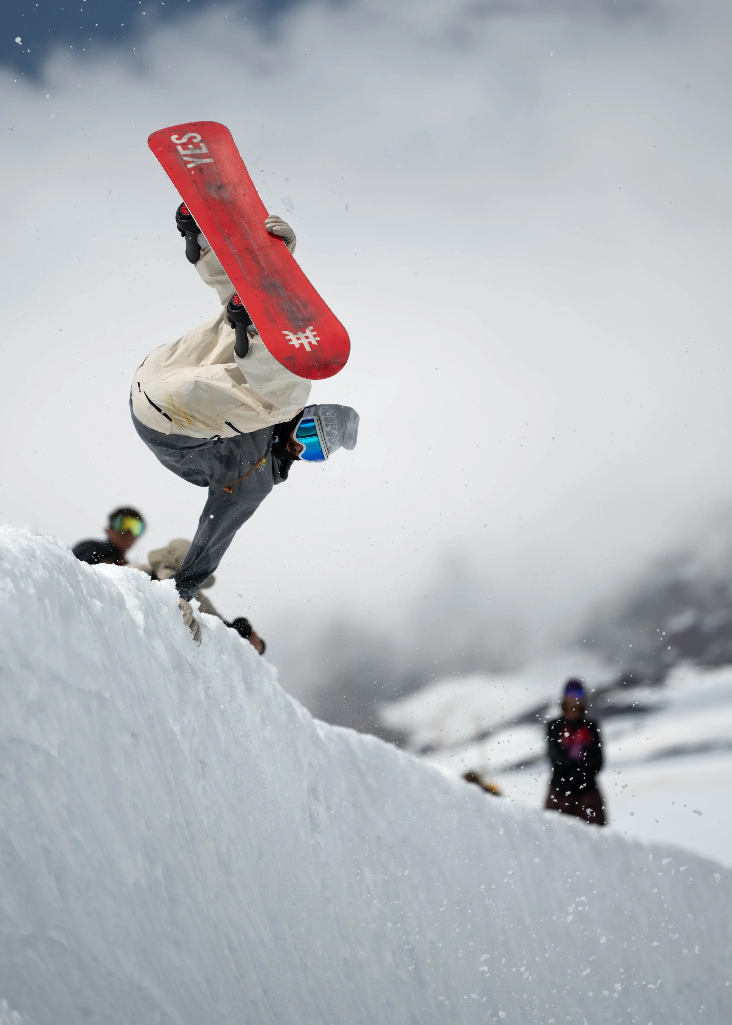 A snowboarder performing a hand plant on a snow-covered slope with other snowboarders in the background.
