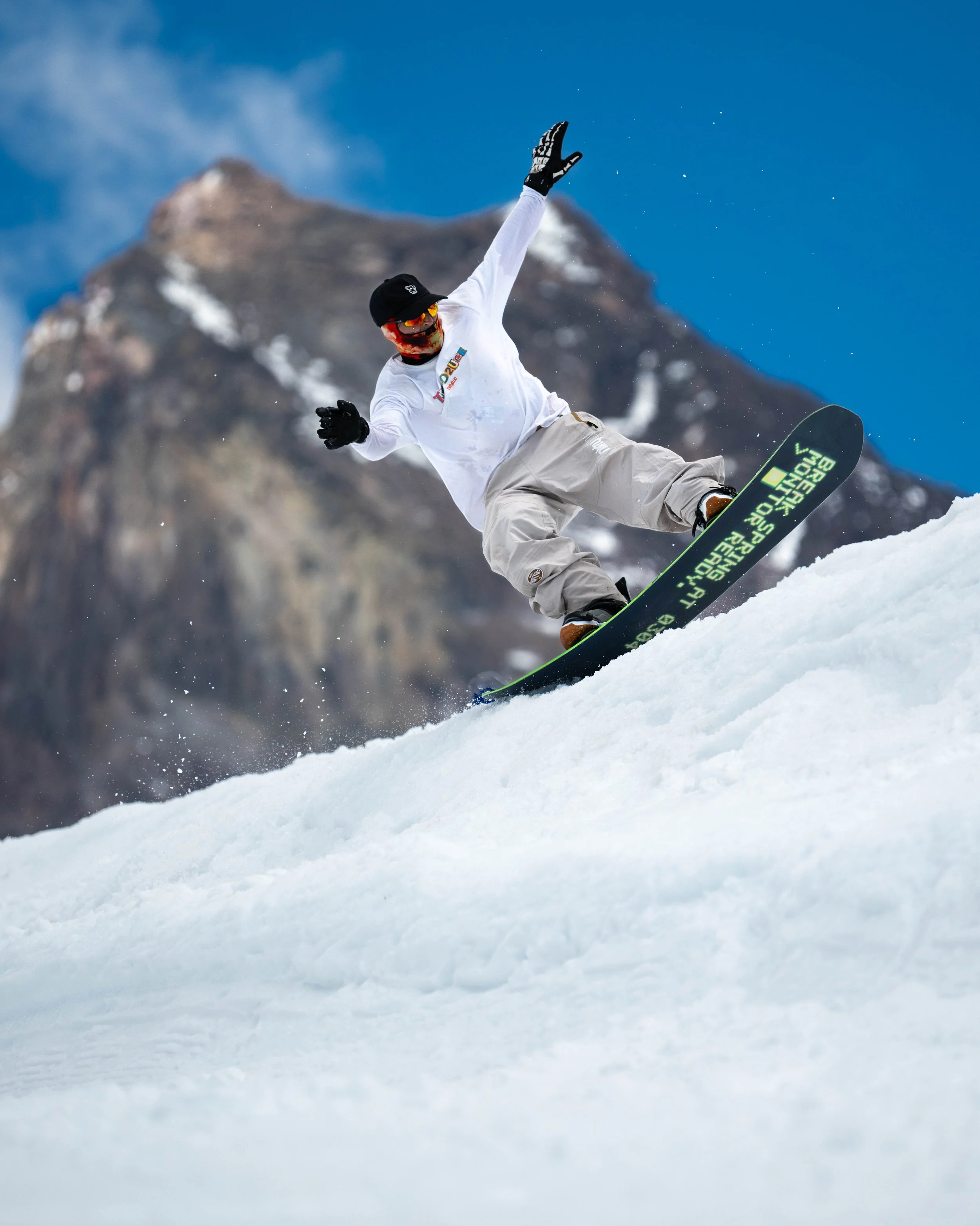 A snowboarder wearing a white shirt, beige pants, black gloves, and a black helmet with sunglasses snowboarding down a snowy slope with a mountain and blue sky in the background.