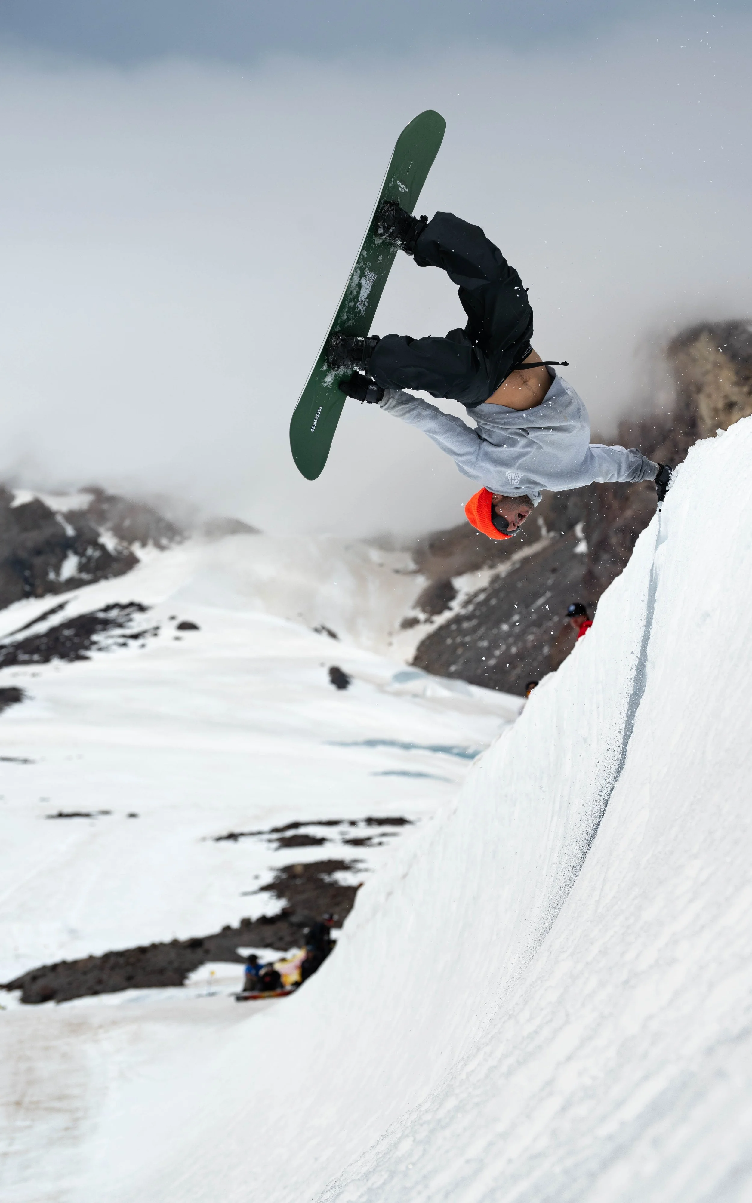 Person performing a hand plant on a snowboard on a snowy mountain slope, wearing a gray jacket, black pants, and an orange beanie.