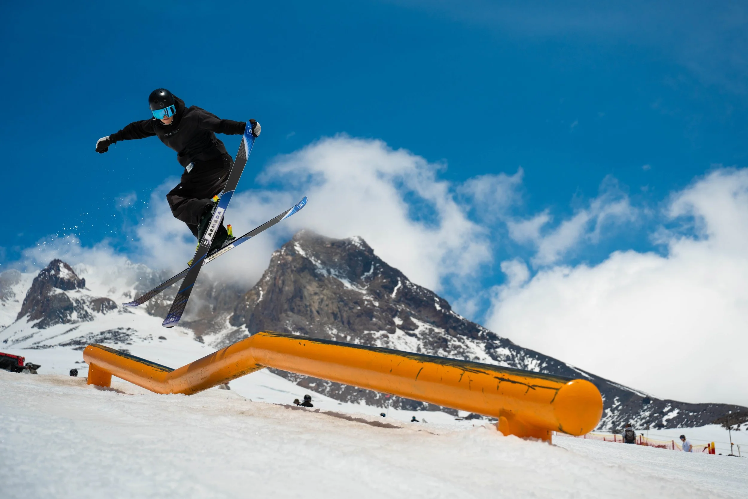 Skier in black gear and a helmet jumps over an orange rail on a snowy mountain slope, with rugged mountains and a partly cloudy sky in the background.