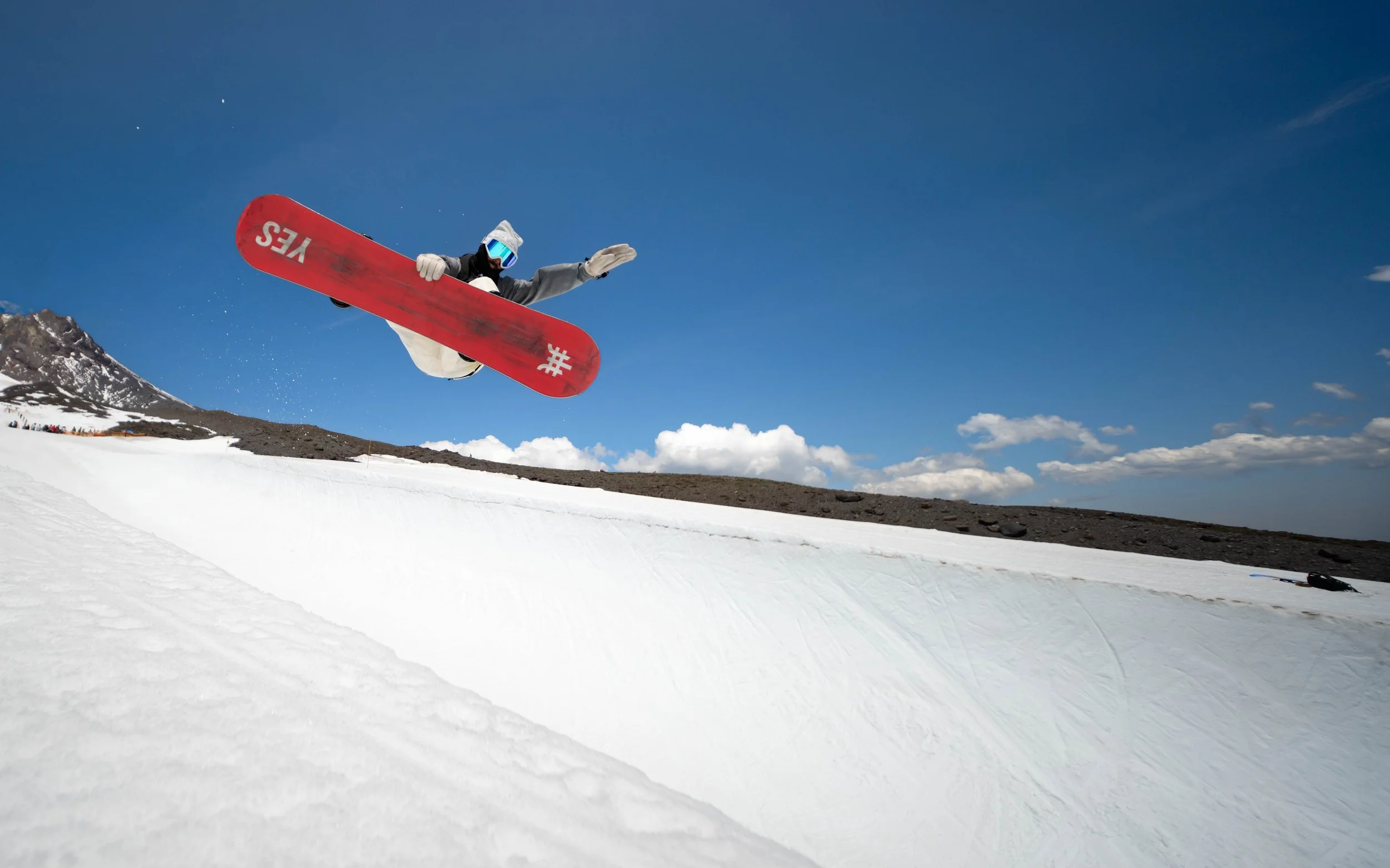 A snowboarder wearing goggles and a hoodie performs a jump on a snowy slope under a blue sky with clouds.