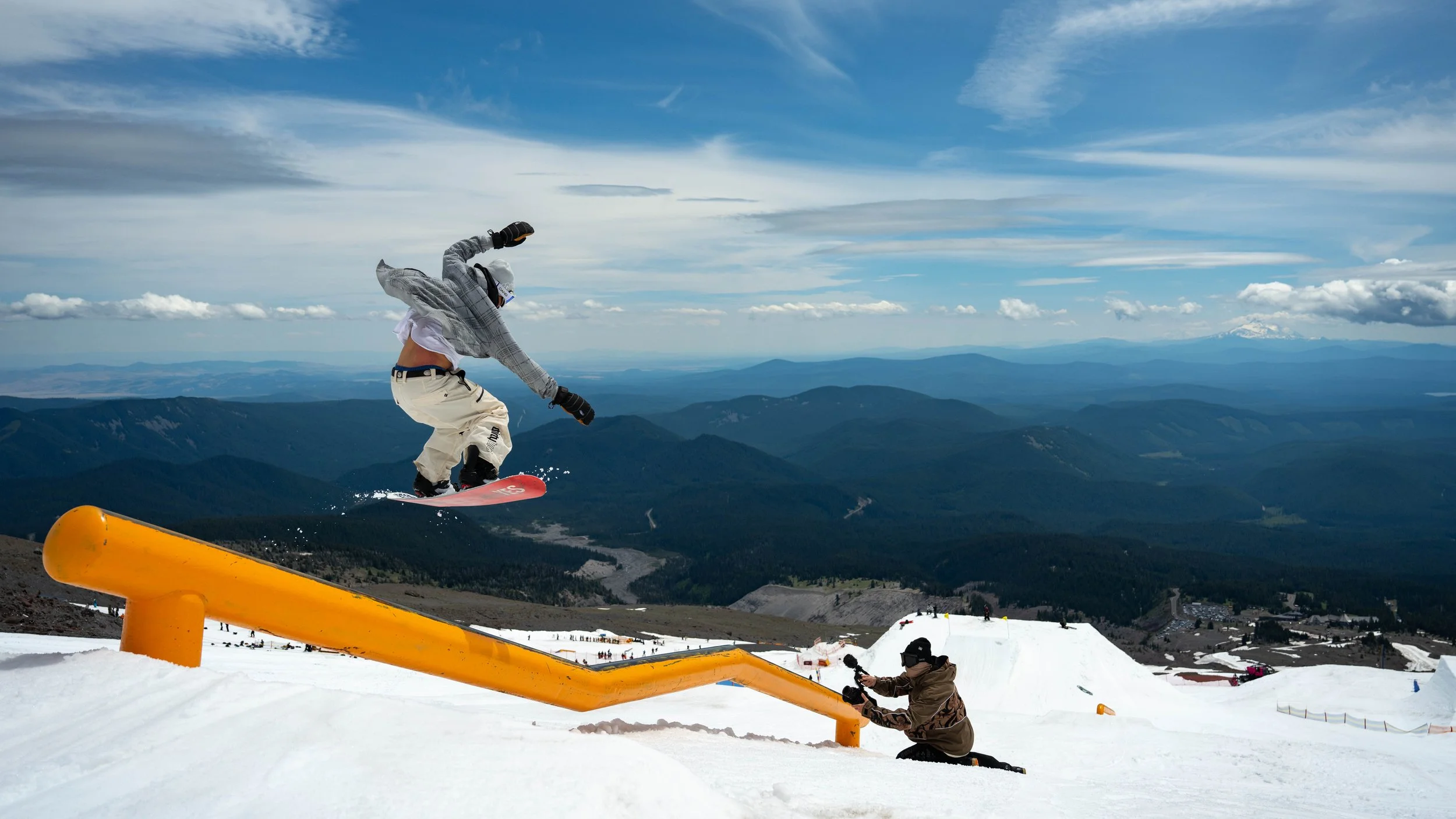 Snowboarder performing a jump over a yellow rail at a snowy ski resort with mountains and blue sky in the background.