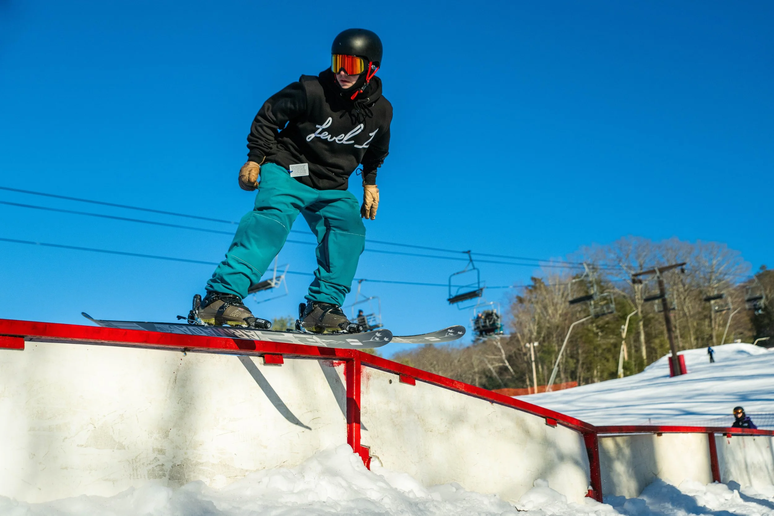 Skier in black hoodie and teal pants performing a rail slide on a snow-covered rail at a ski resort.