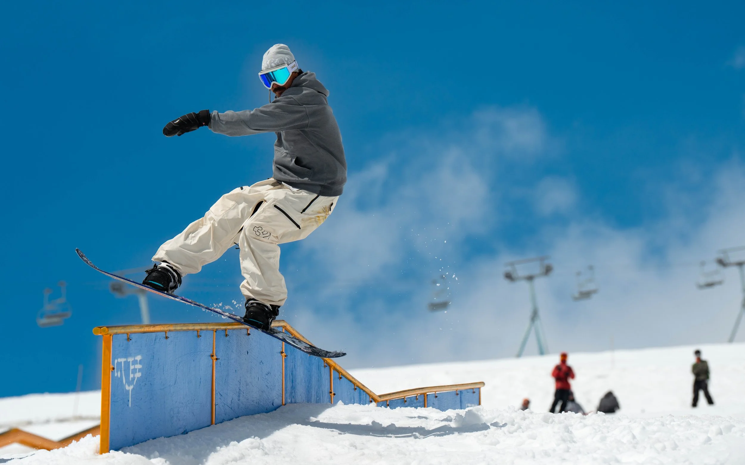 A snowboarder in a gray jacket, white pants, black gloves, and a gray beanie performs a trick on a blue rail at a snowy ski resort with chairlifts and other skier spectators in the background under a clear blue sky.