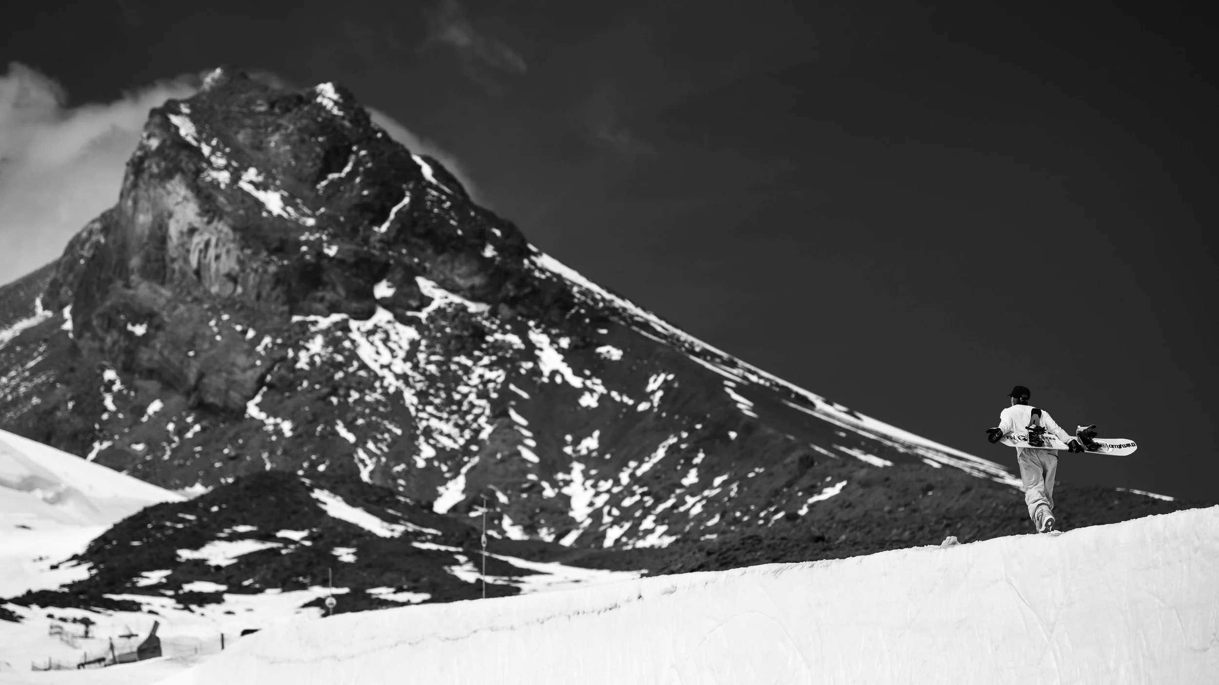 A person in snowboarding gear carrying a snowboard walking on snow with a large mountain in the background, black and white photo.