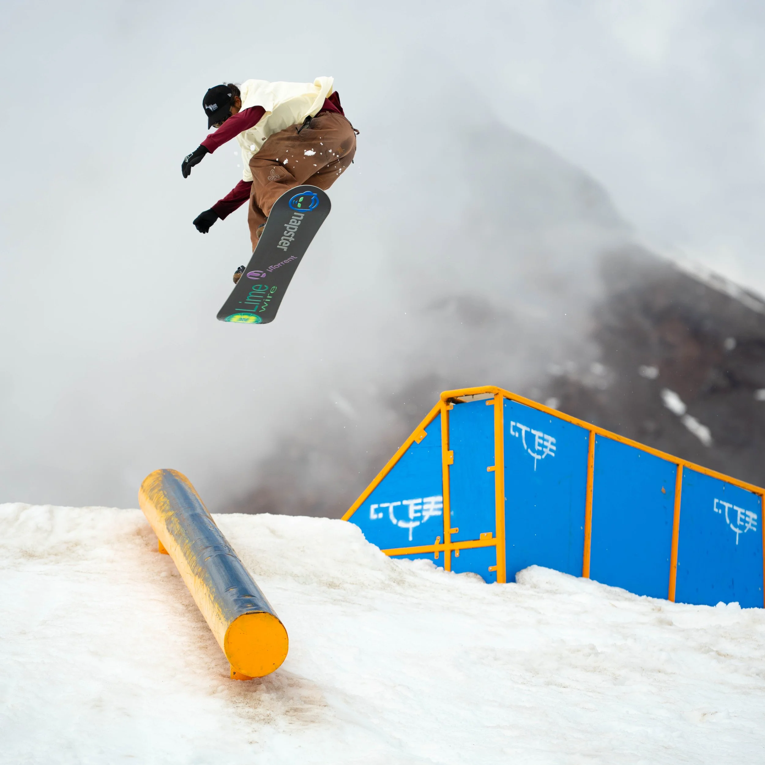 A snowboarder in mid-air performing a jump over a snow ramp at a ski resort, with mountain and cloudy sky in the background.