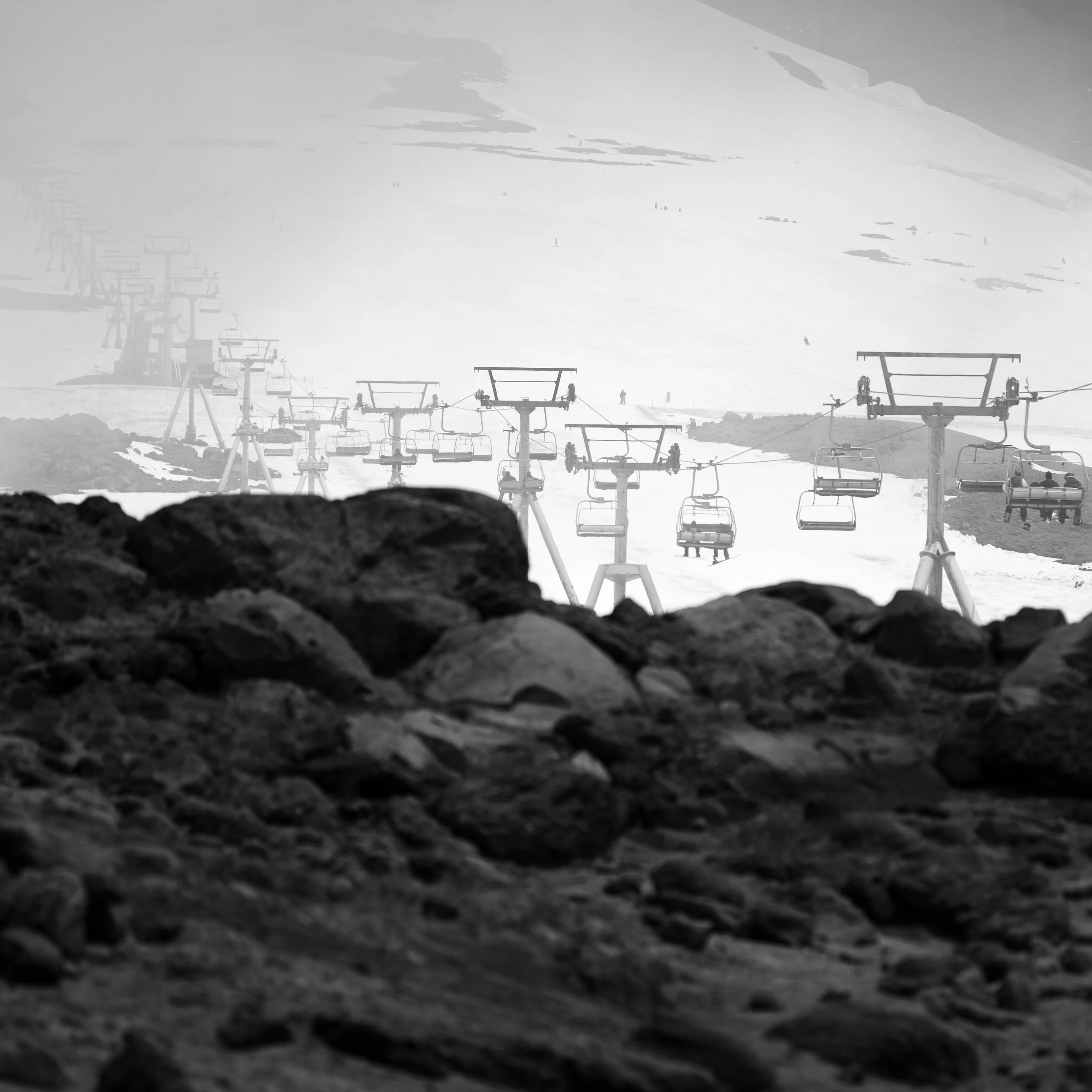Black and white photo of ski lift chairs on a snowy mountain, with rocky foreground and cloudy sky.