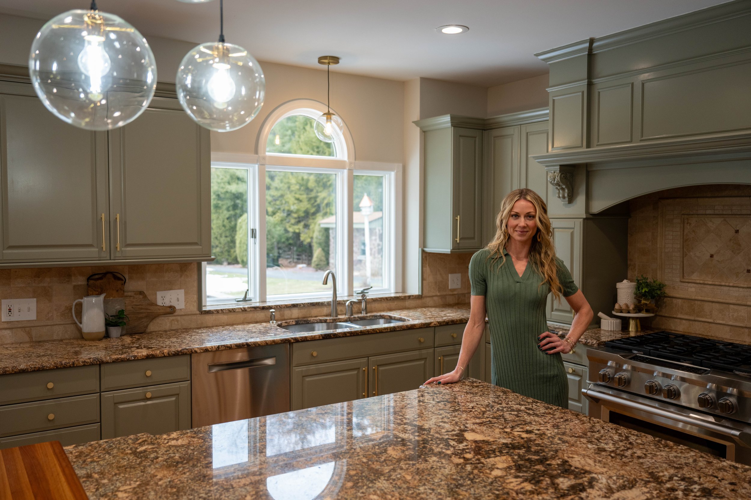 A woman with long blonde hair wearing a green dress standing in a spacious kitchen with granite countertops and green cabinets, near a large window with a view of outdoor greenery.