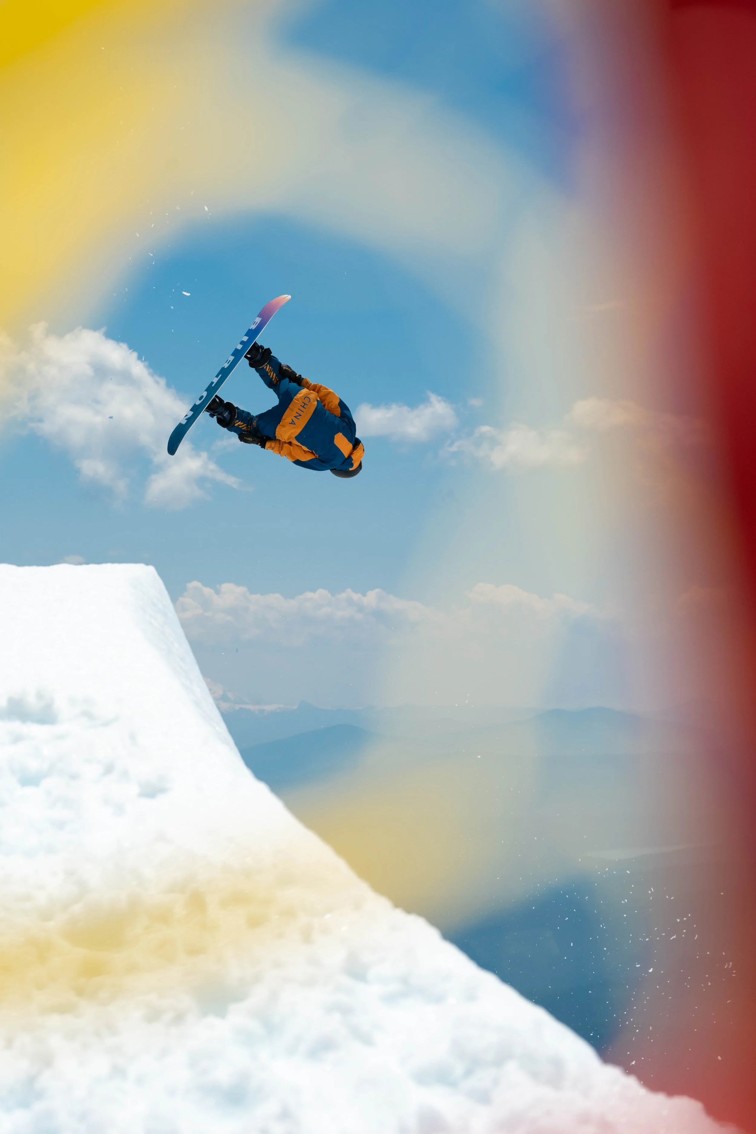 A snowboarder wearing a blue and orange jacket performing an aerial trick above a snow ramp on a clear, sunny day.