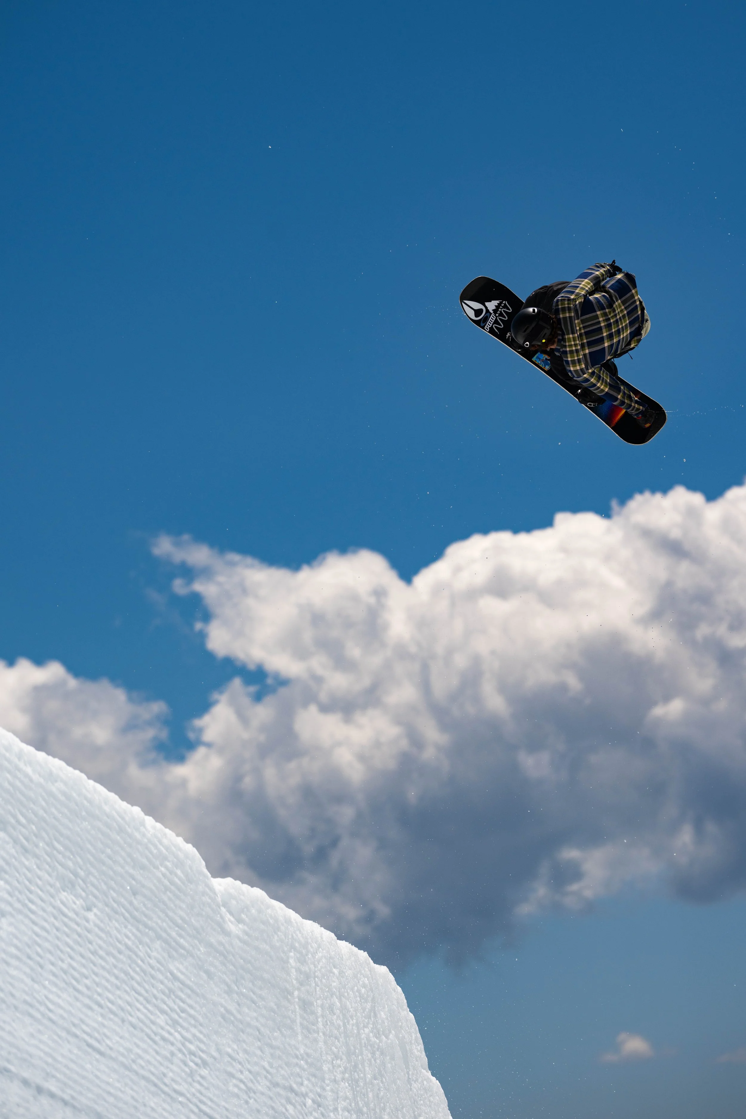 A snowboarder in a plaid jacket and helmet performing a jump in mid-air against a blue sky with clouds and snow on a winter slope.