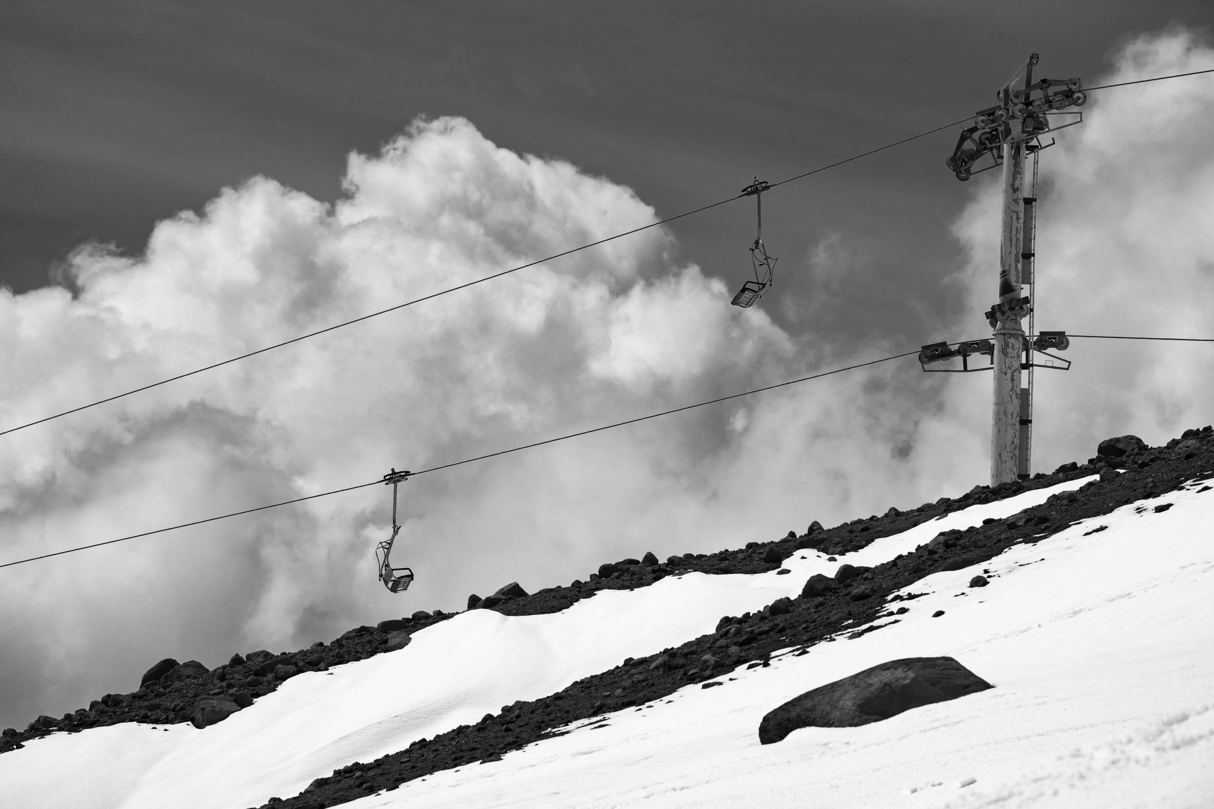Empty ski lift chairs hanging from a cable on a snow-covered mountain slope with rocks, against a cloudy sky.