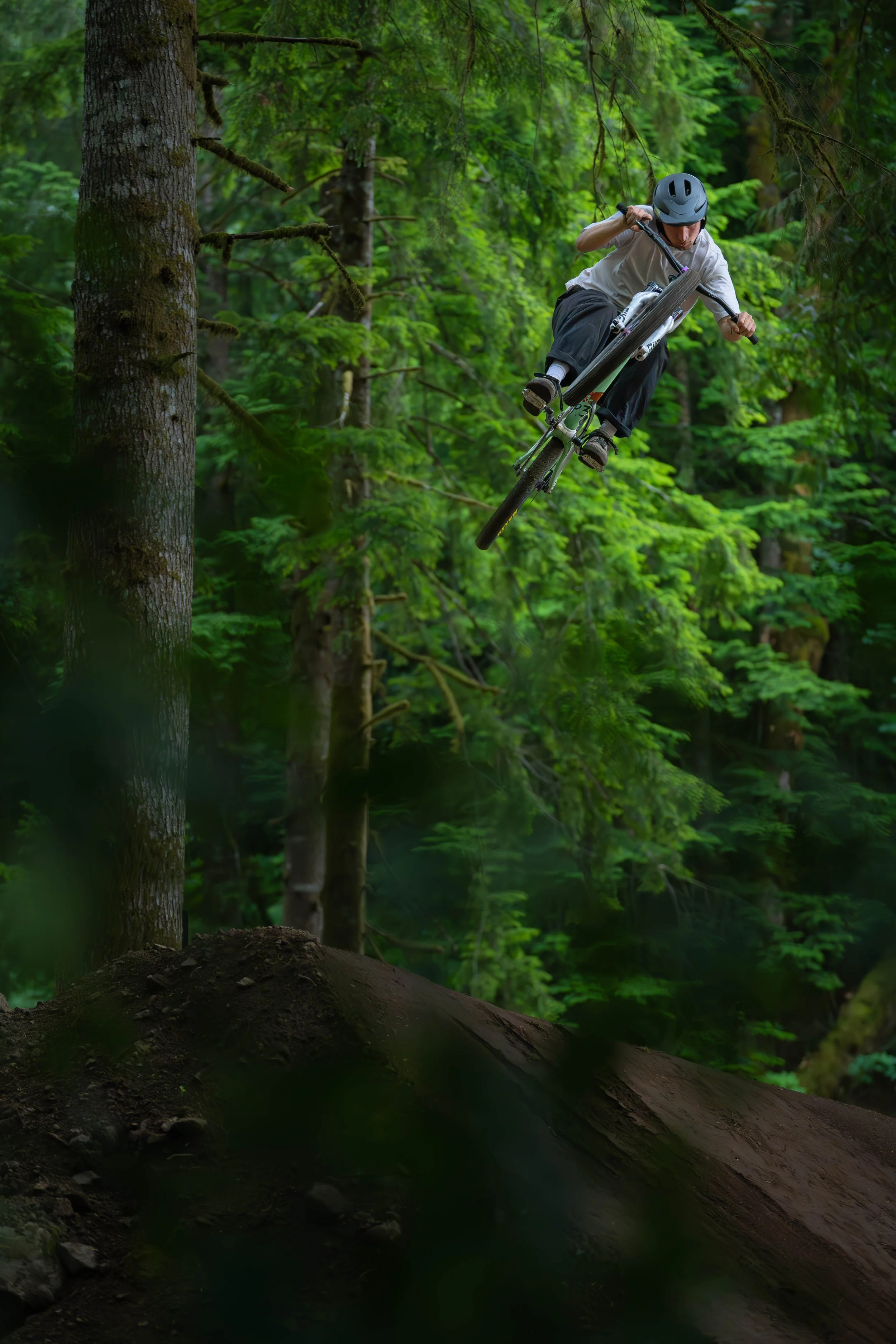 A person wearing a helmet riding a mountain bike in mid-air above a dirt trail in a dense green forest.