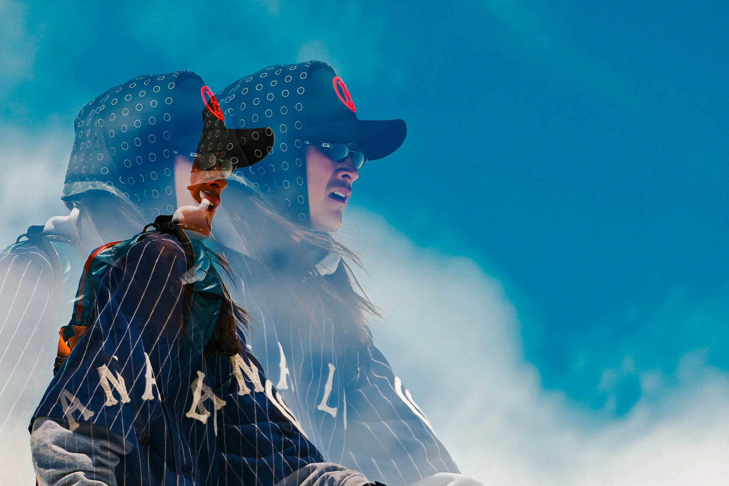 Two people wearing blue helmets and jackets with the word 'VAIL' in white, standing against a bright blue sky with some clouds.