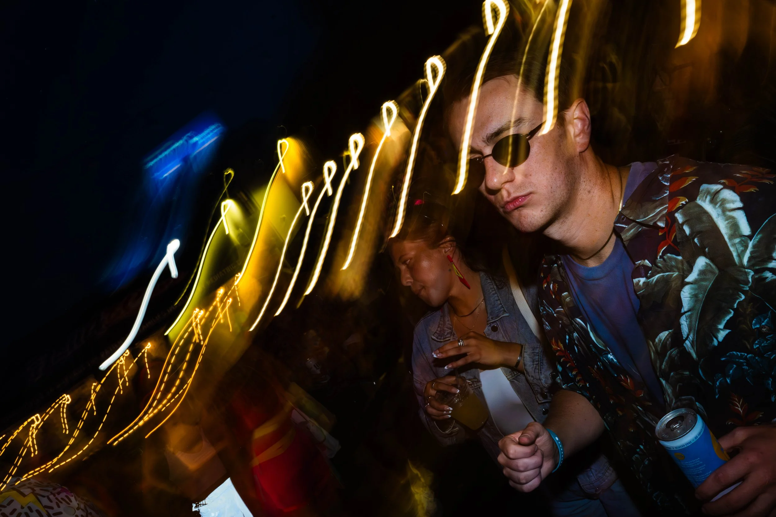 A man wearing sunglasses and a printed shirt holding a canned beverage at a nighttime event, with blurred yellow and white string lights creating streaks across the dark sky.