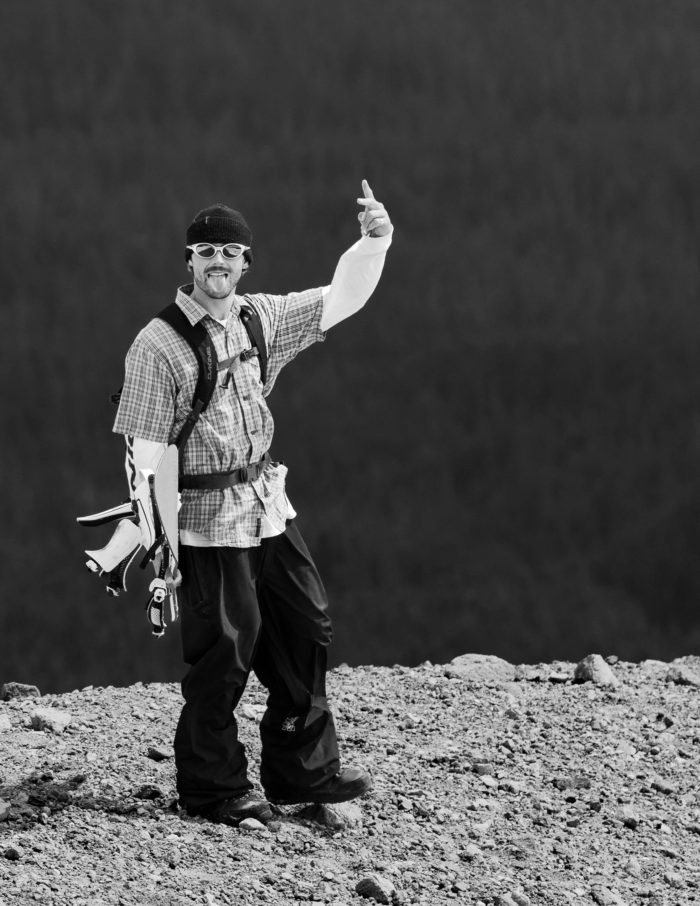 A man standing on rocky terrain, wearing sunglasses, a beanie, a plaid shirt, and carrying climbing gear, making a gesture with his hand and sticking his tongue out.
