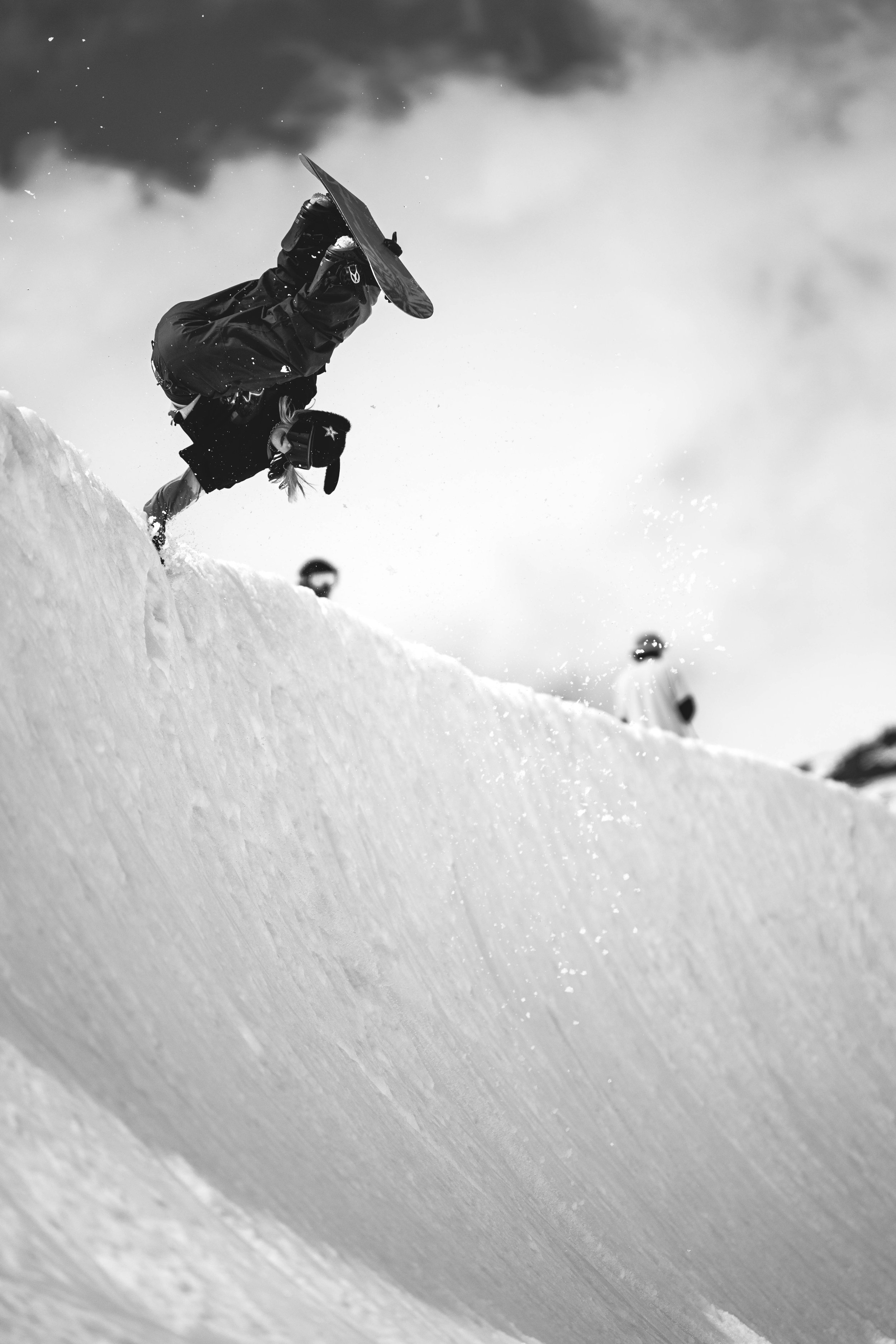 A snowboarder performing a hand plant on a snowy incline, captured from a low angle in black and white, with two other snowboards in the background.