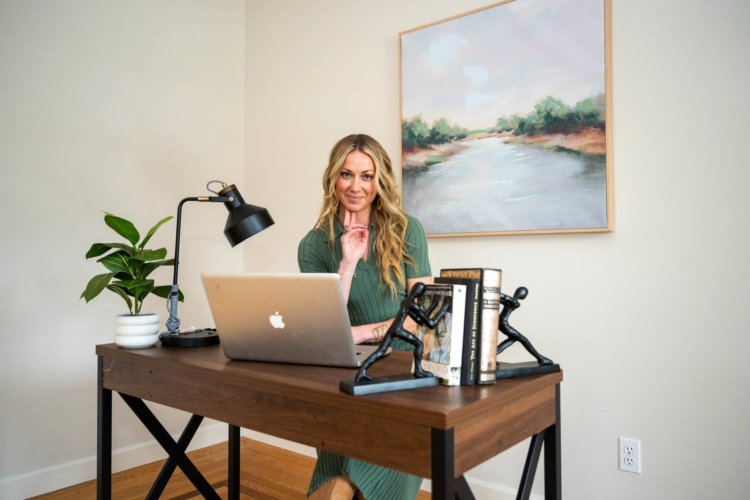 A woman with long blonde hair, sitting at a wooden desk with a laptop, placed in a room with a painting of a river on the wall. There is a potted plant and decorative sculptures of people on the desk.