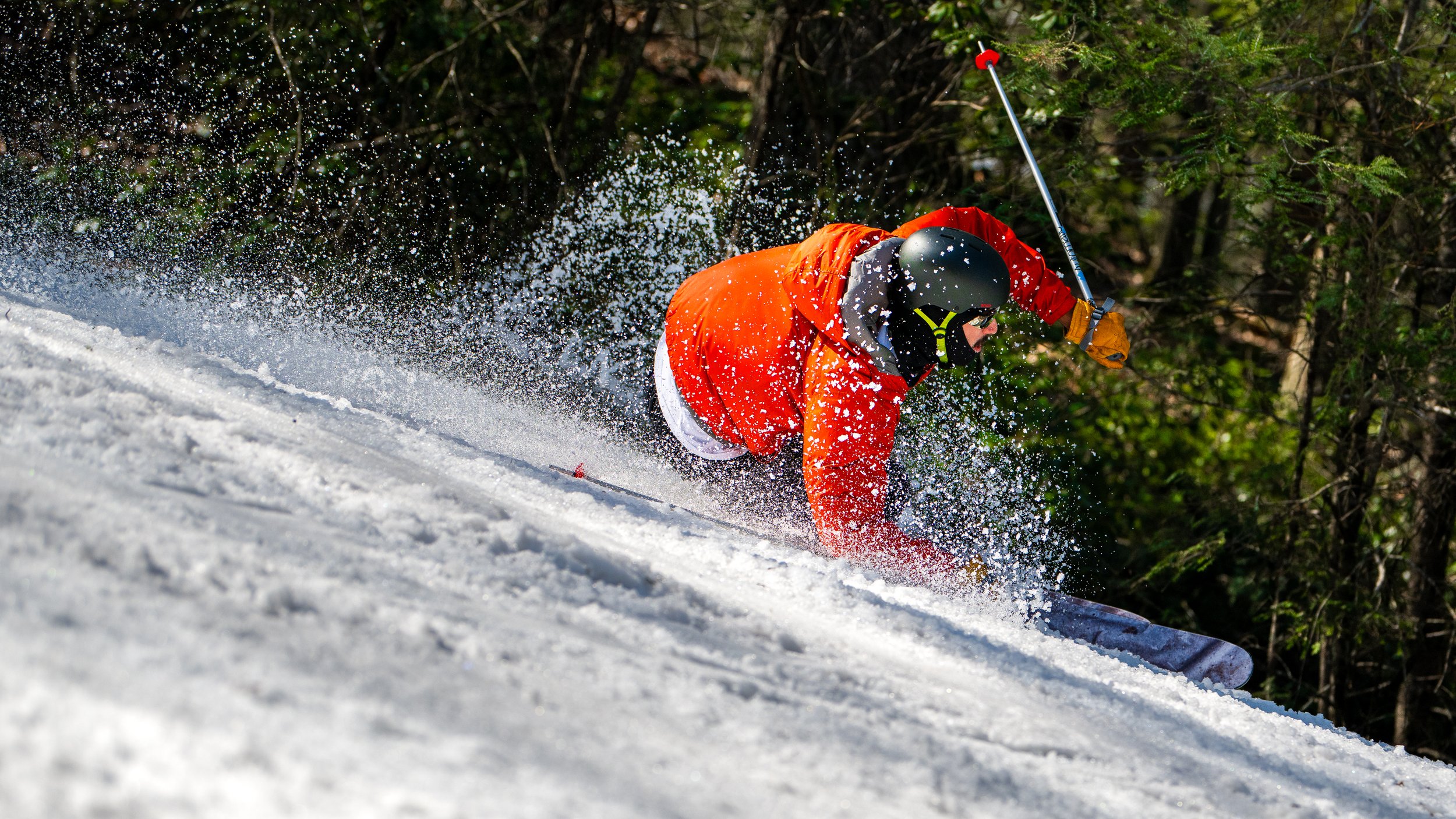 A skier in an orange jacket and black helmet falling forward on a snow-covered slope surrounded by green trees.