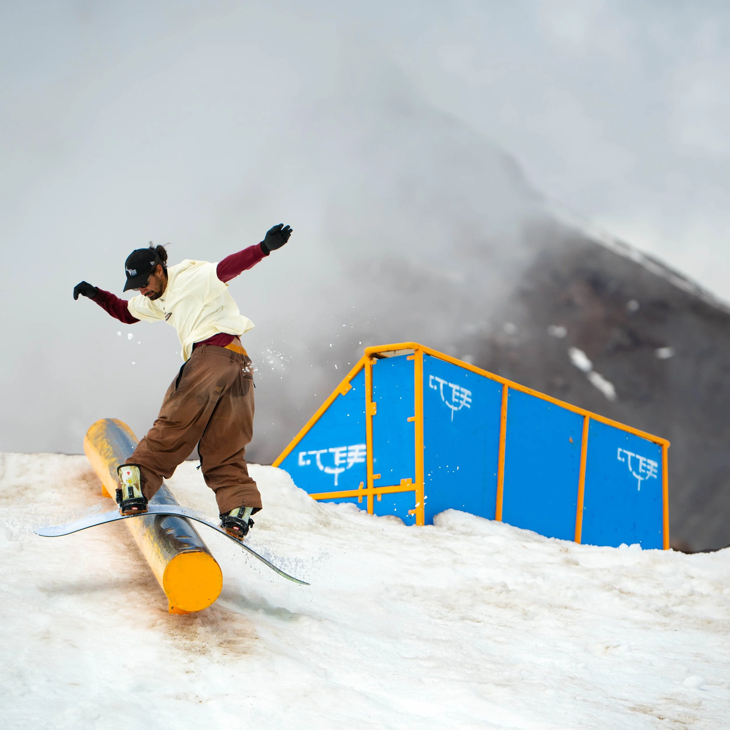 Skateboarder performing a trick on a rail in a snowy mountain setting under cloudy sky.