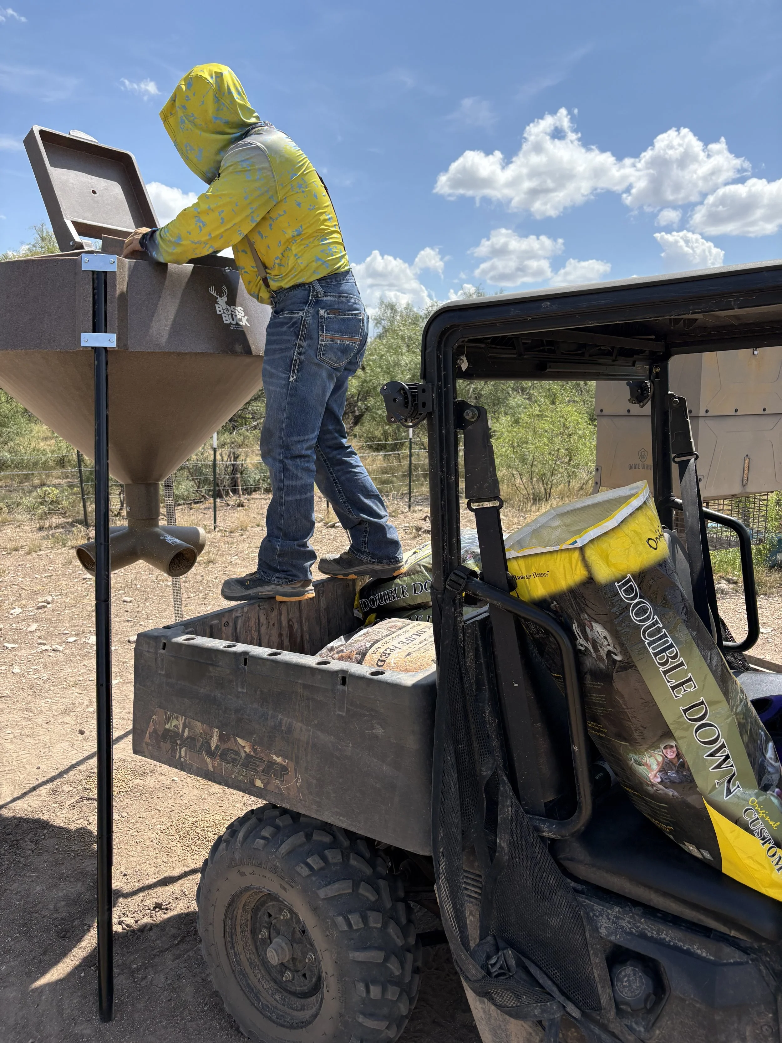 A person standing on a utility vehicle bed, loading bags of deer feed into a large deer feeder in an outdoor, rural setting with a blue sky and scattered clouds.