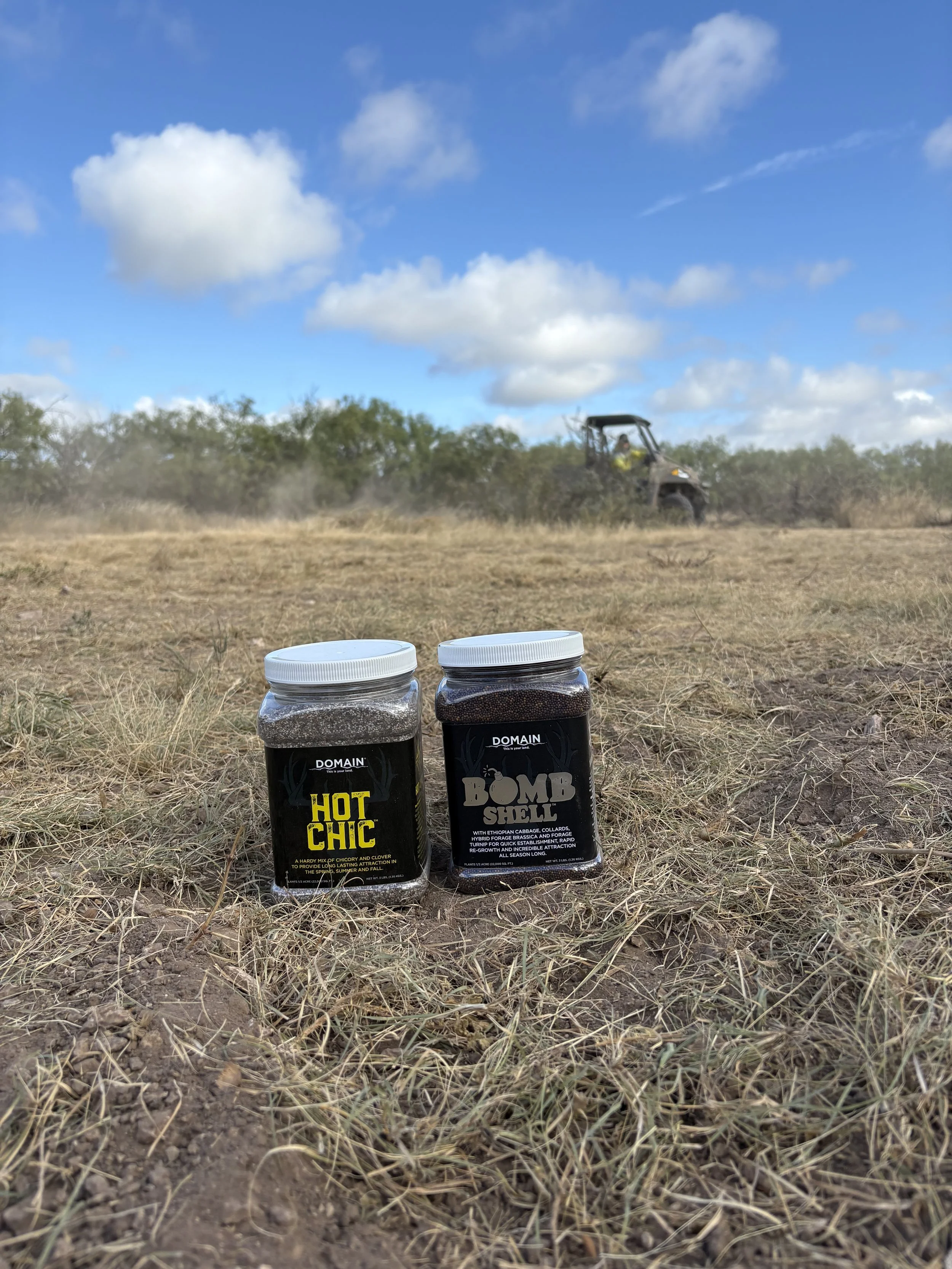Two jars of gardening products labeled 'Hot Chic' and 'Bomb Shell' on dry ground with sparse grass, and a UTV vehicle working on the open field in the background under a bright blue sky with some clouds.