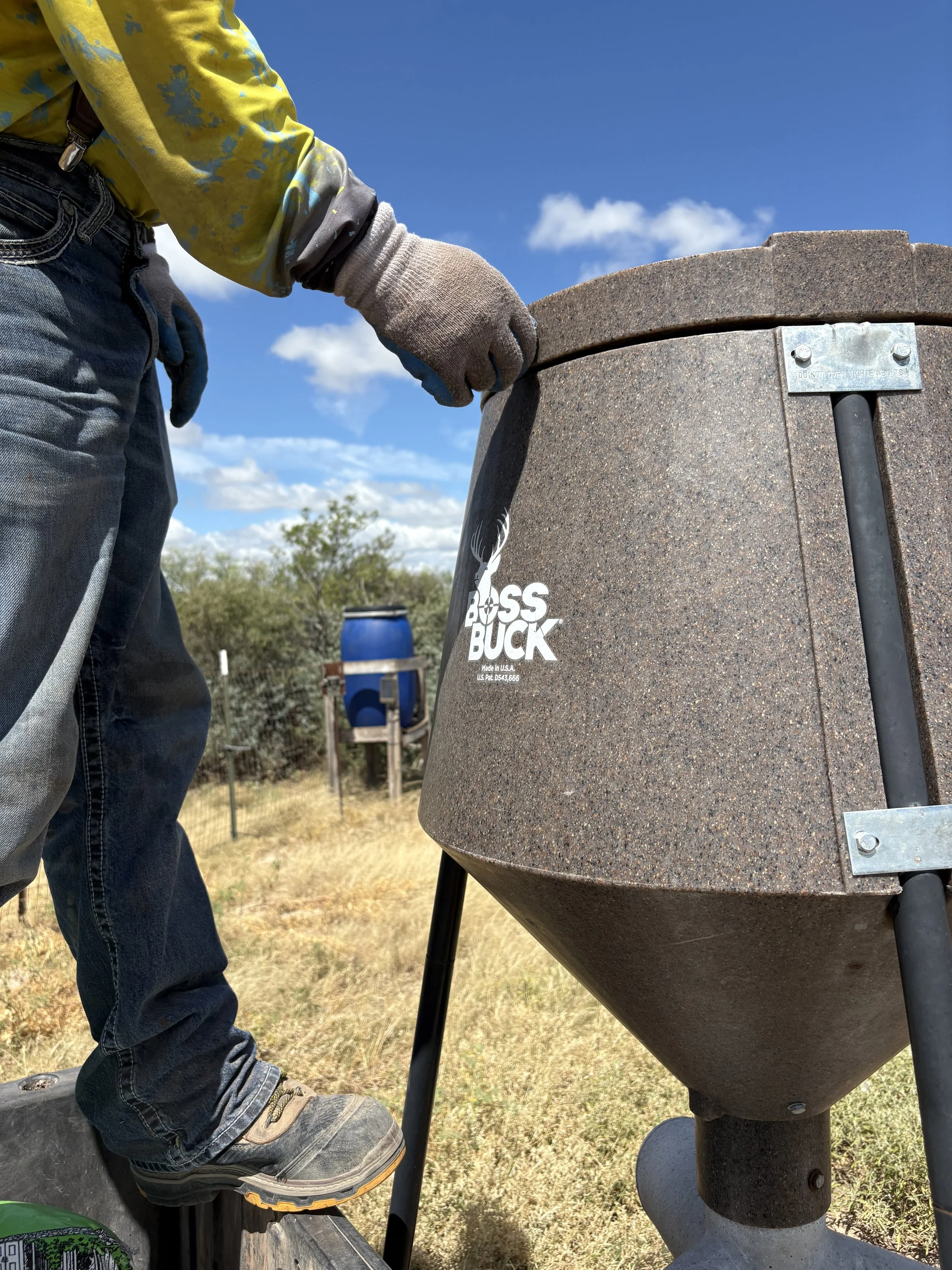 A person wearing gloves and jeans is filling a large, brown, cone-shaped black powder or grain hopper with a blue container in the background, outdoors on a sunny day.